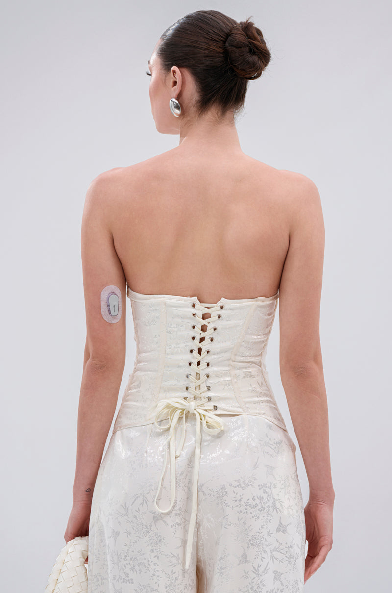 A woman with dark hair in a bun, facing away from the camera, wears the DAISY DOVE JACQUARD CORSET with structural boning and white patterned pants. She has a medical device on her upper arm and holds a woven white clutch.