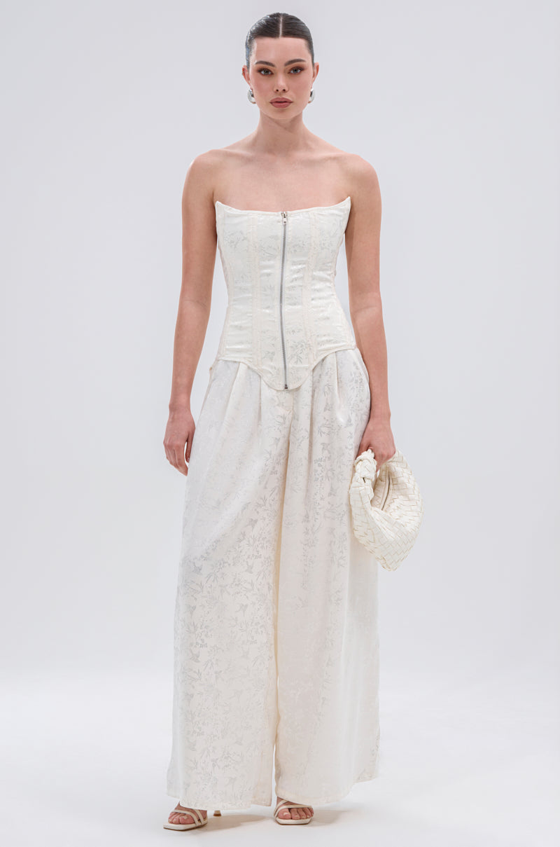 A woman poses against a plain light background wearing the DAISY DOVE JACQUARD CORSET, paired with wide-leg white patterned pants, a textured white clutch, and open-toe sandals.