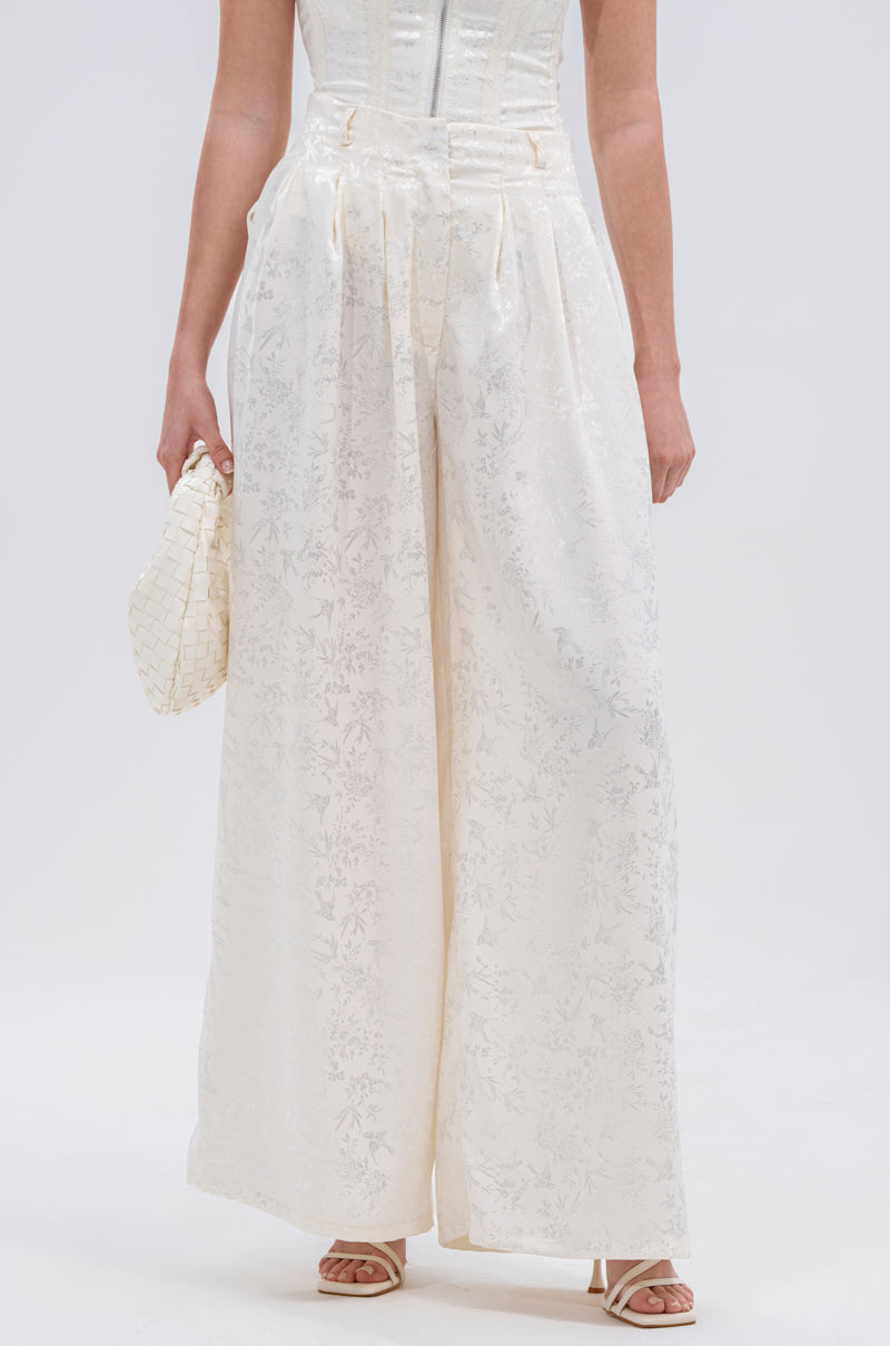 Wearing DAISY DOVE JACQUARD TROUSER featuring a high waist, wide-leg cut, and subtle floral pattern, paired with a textured white clutch and open-toed heels. The top of the outfit is partially visible.