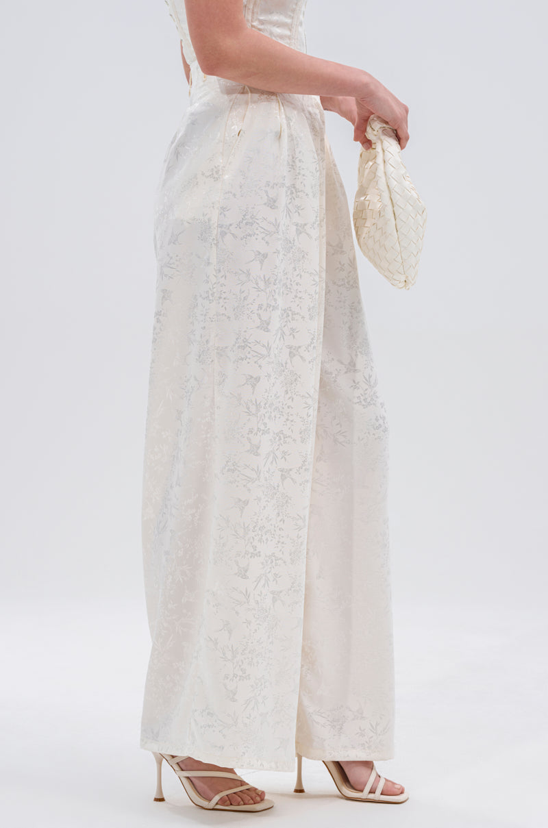 A woman in profile wears DAISY DOVE JACQUARD TROUSER with a wide-leg silhouette, open-toe heels, and holds a small woven clutch. The floral pattern pops against the plain white background.