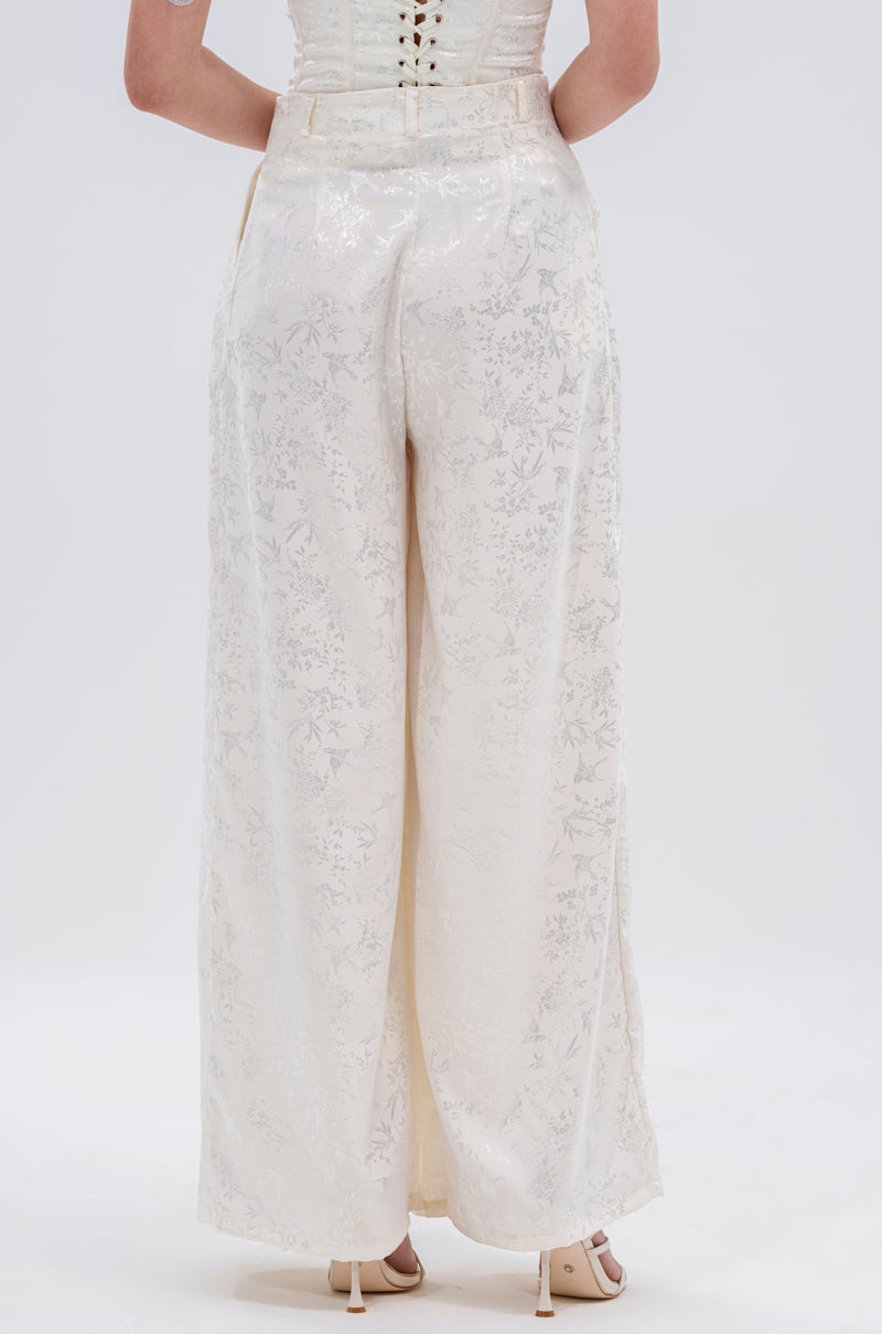 A person is seen from the back wearing DAISY DOVE JACQUARD TROUSER featuring a high waist, wide-leg silhouette, and subtle floral pattern, paired with beige heeled sandals, set against a plain light background.