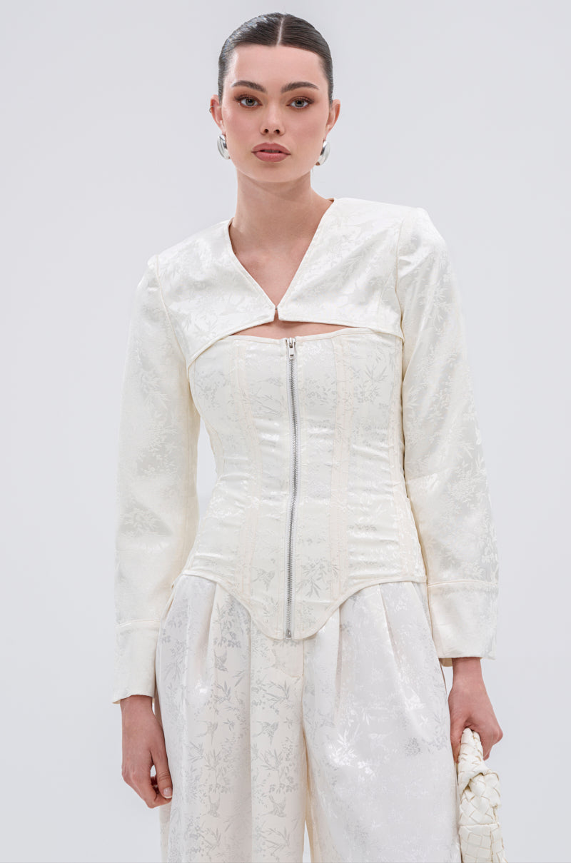Wearing the DAISY DOVE JACQUARD BLAZER with a zip-up corset front and wide-leg pants, a woman stands against a light background holding a small white textured bag, radiating vintage-inspired elegance.