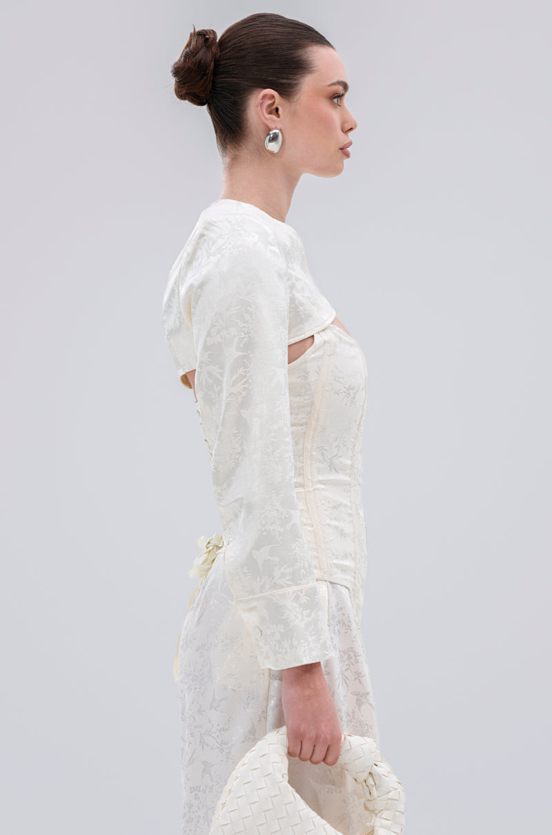 A woman with dark hair in a sleek bun wears the DAISY DOVE JACQUARD BLAZER, an elegant, textured white piece with long sleeves. Pictured in profile, she holds a woven white clutch against a plain light backdrop, exuding vintage-inspired style.