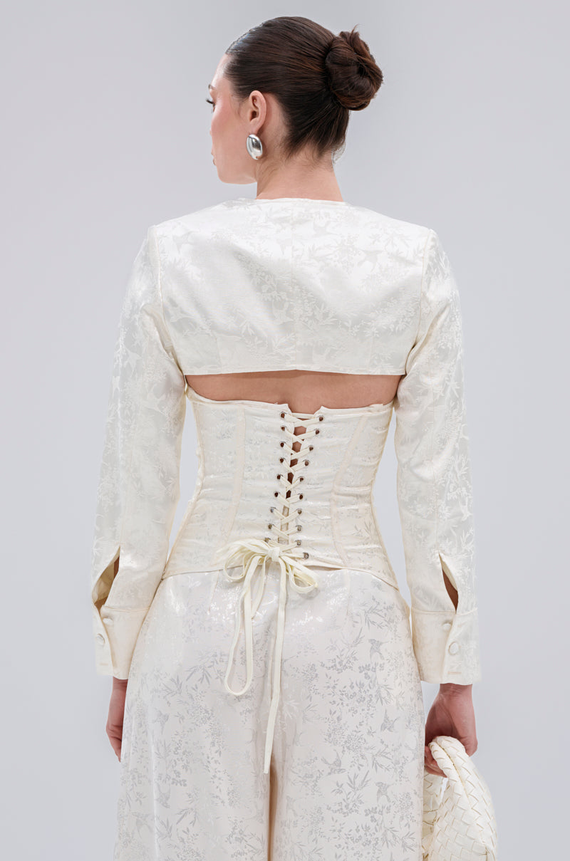 A woman with dark hair in a sleek bun wears the elegant DAISY DOVE JACQUARD BLAZER, a patterned white satin jacquard piece with a lace-up corset-style back. She holds a textured white clutch and faces away from the camera.