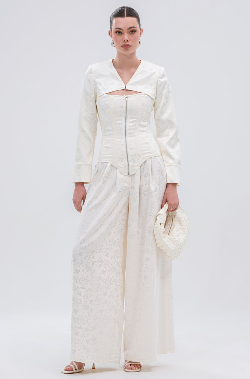 A woman poses against a light background wearing the DAISY DOVE JACQUARD BLAZER—an elegant, textured white satin jacquard piece styled with wide-leg pants, a fitted top, sandals, a woven white clutch, and her hair pulled back.
