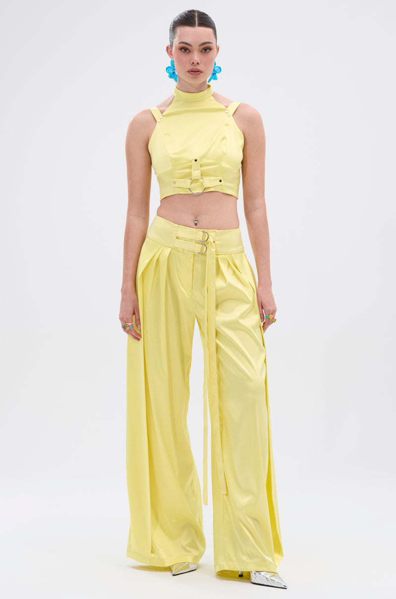 A woman faces forward in a bright yellow sleeveless crop top with buckles and ELECTRIC SATIN TROUSER, both in high-shine satin. She wears her hair slicked back, blue statement earrings, and silver pointed shoes against a plain backdrop.