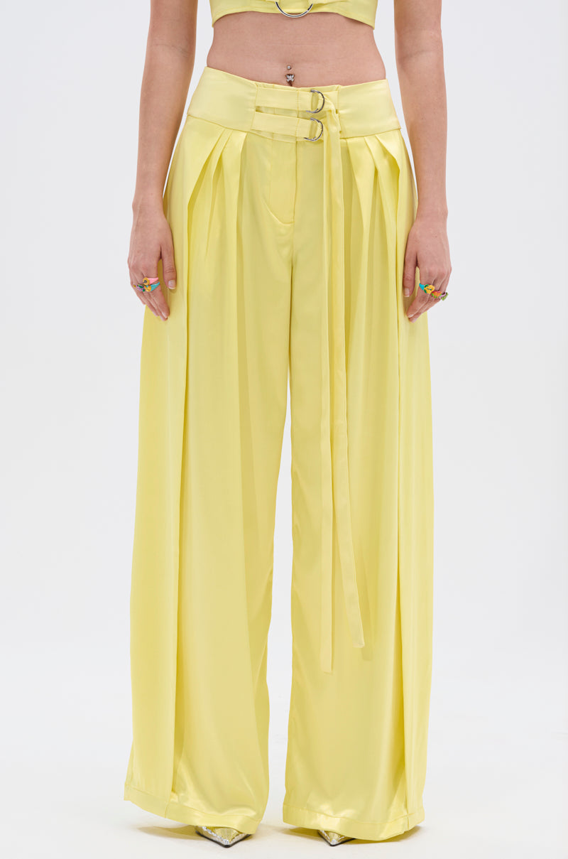A person wears the ELECTRIC SATIN TROUSER—high-waisted, wide-leg pants with pleats and an adjustable belt—plus pointed shoes. The glossy satin fabric shimmers as only their upper body is partially shown against a white backdrop.