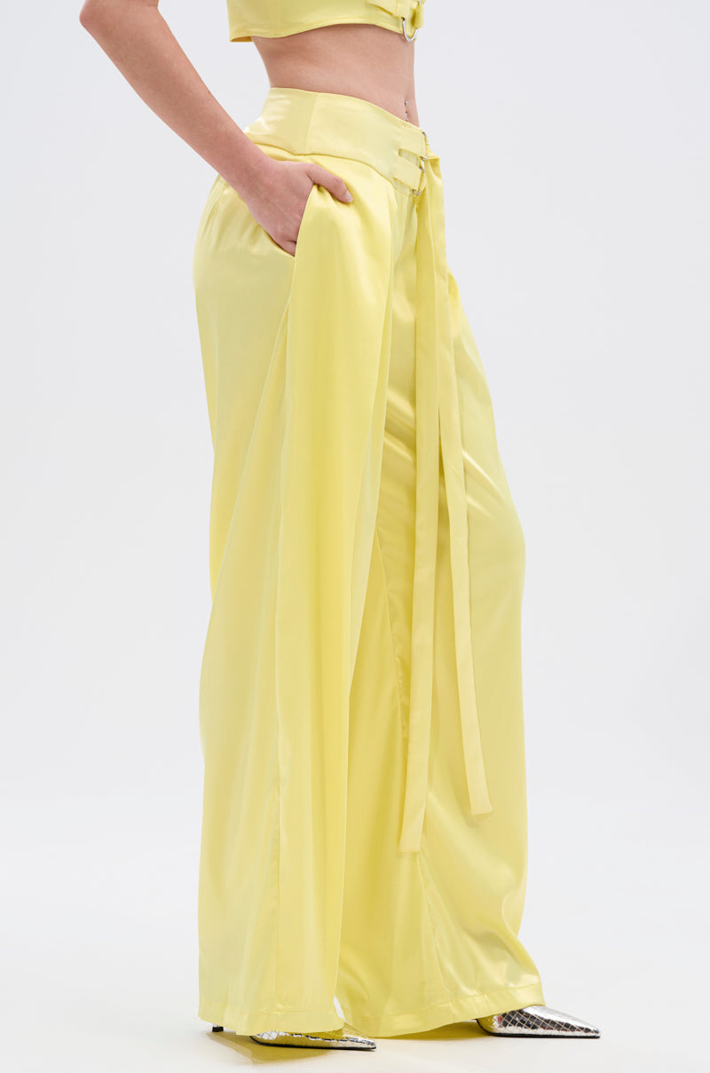 Wearing the yellow ELECTRIC SATIN TROUSER, high-waisted and wide-leg in glossy satin with pockets and adjustable belt accents, paired with a matching crop top; trousers mostly cover silver-detailed pointed-toe shoes. Plain white background.