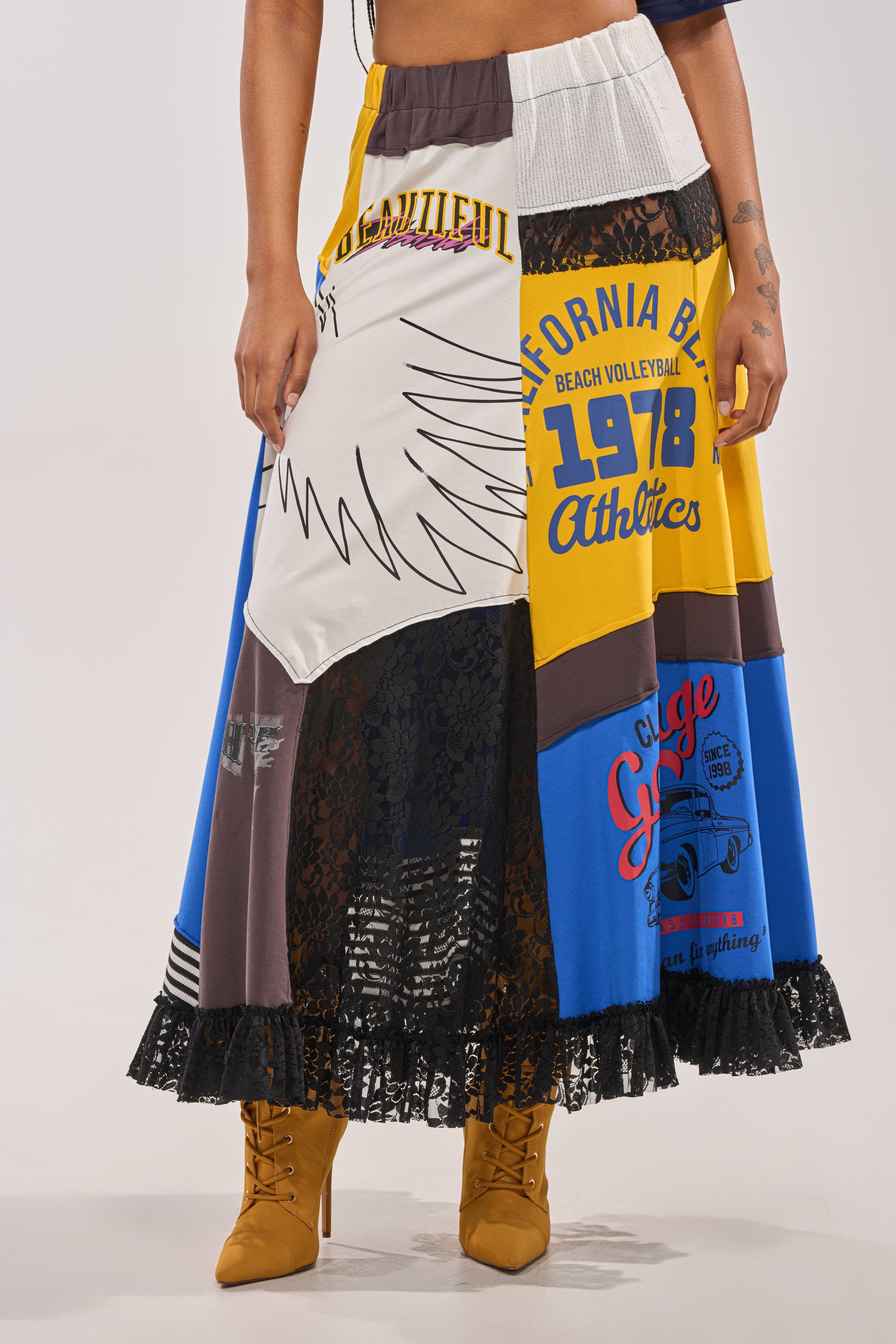 A woman wears the CRAZY FOR YOU MAXI SKIRT featuring colorful graphics, lace panels, and text, paired with tan lace-up heeled boots. Only the lower half of her body is visible. 0