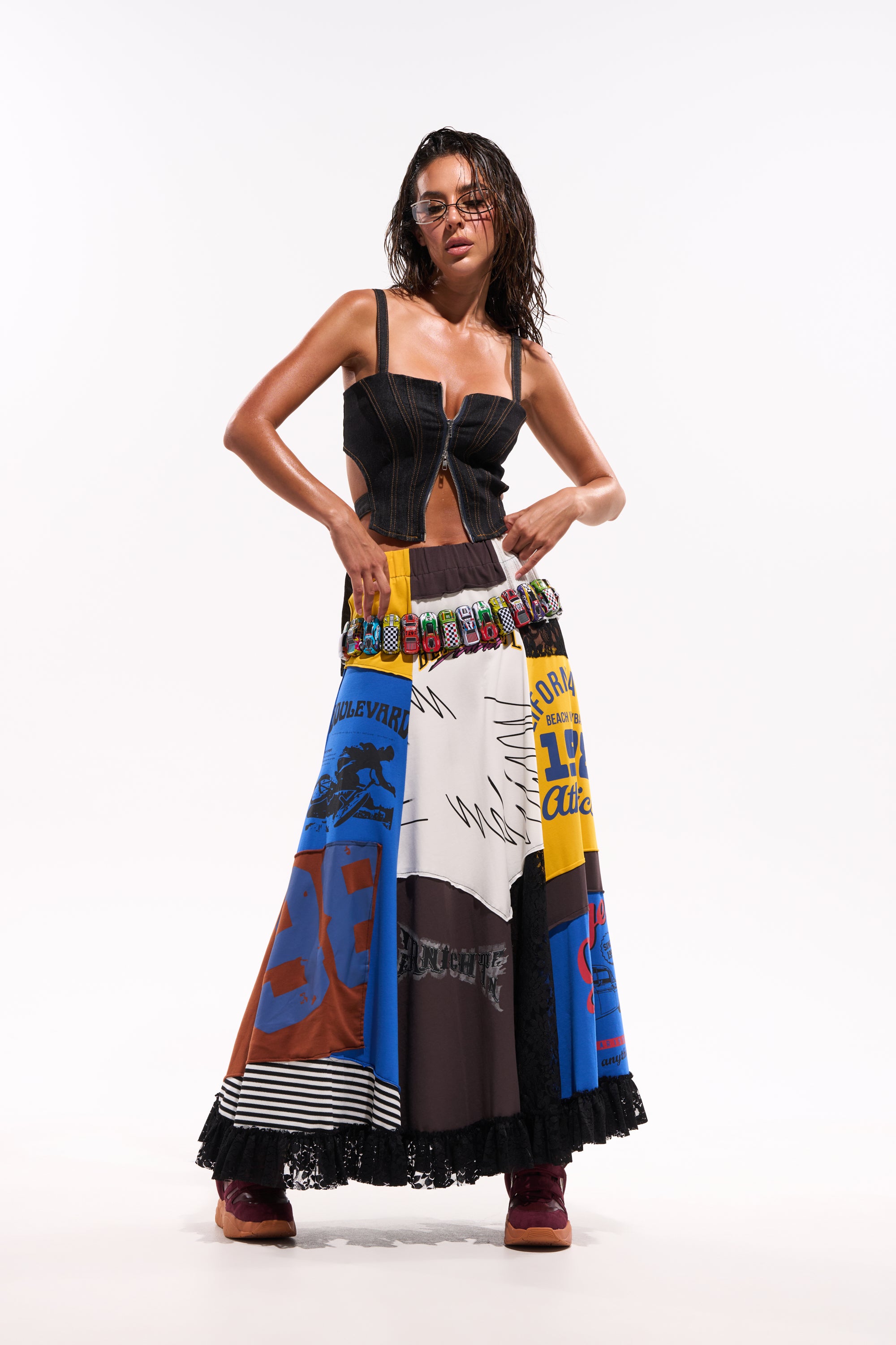 A woman wearing glasses poses confidently in a black corset top and the CRAZY FOR YOU MAXI SKIRT, featuring sporty patchwork graphics and numbers. She stands against a white background, hands on her waist, showcasing bold game day style. 2
