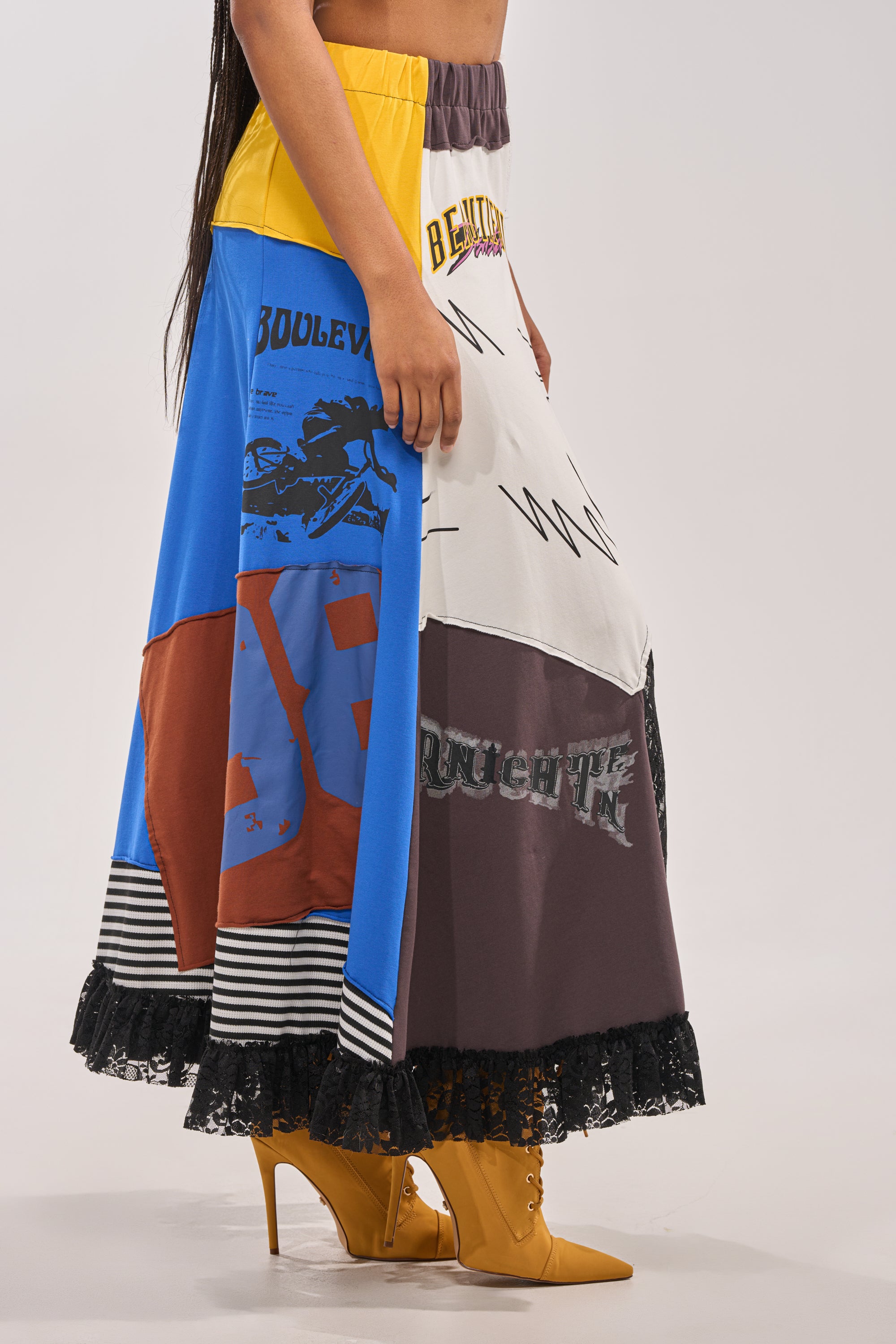 A person wearing the CRAZY FOR YOU MAXI SKIRT, featuring graphic patchwork and lace trim at the hem, styled with yellow heeled boots. The upper body is out of frame, highlighting the vibrant maxi skirt and bold footwear. 3