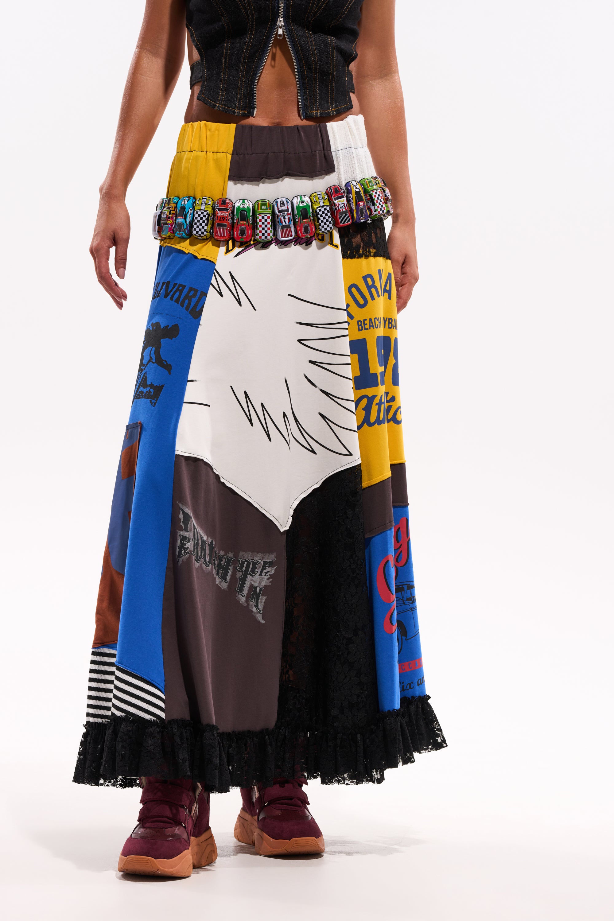 A person wears the CRAZY FOR YOU MAXI SKIRT with graphic fabrics, accessorized with a belt of toy cars, maroon sneakers, and a black crop top. The photo is set against a plain white background. 1