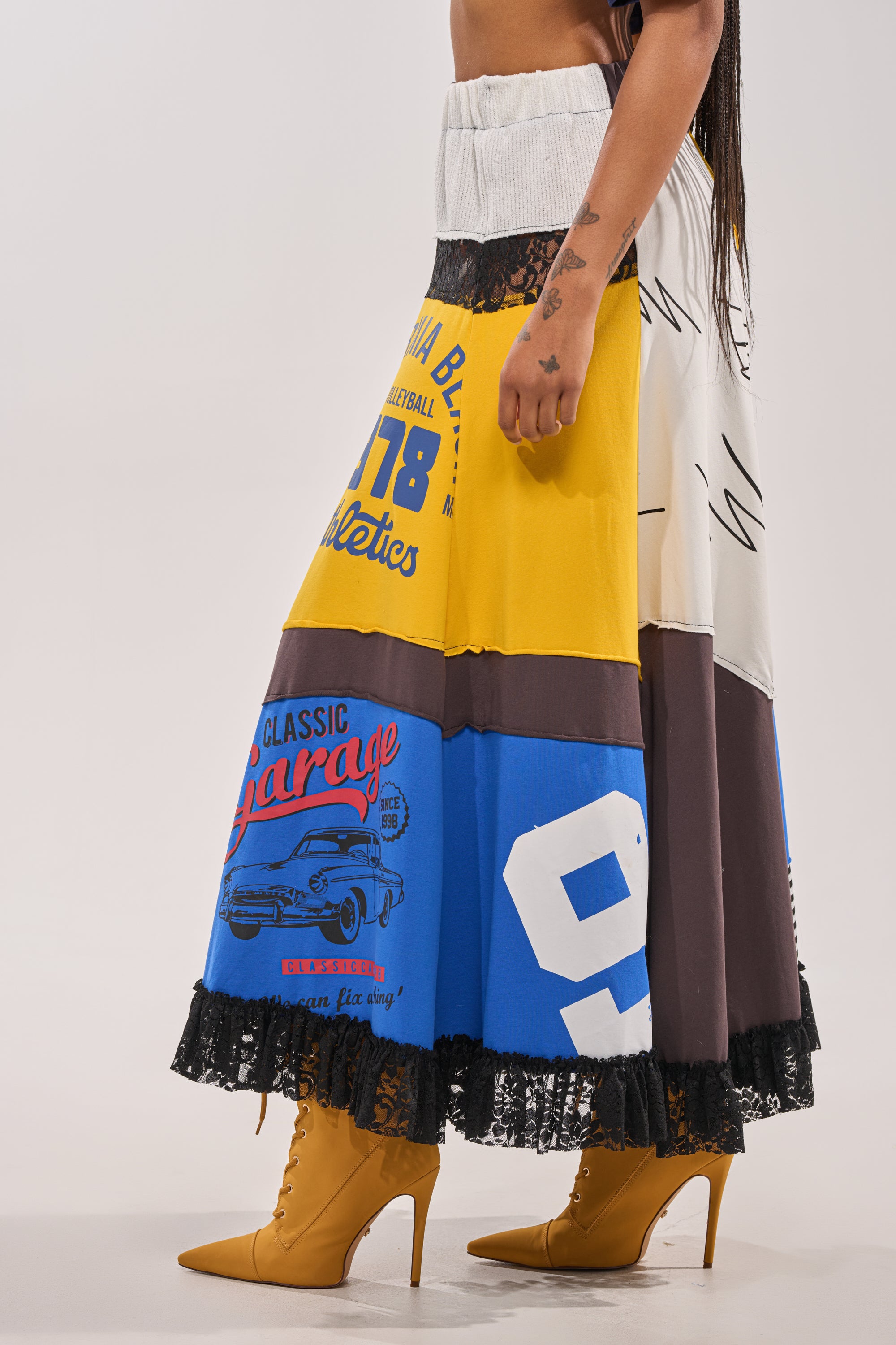 The lower body is shown wearing the CRAZY FOR YOU MAXI SKIRT, a sporty mixed-print maxi with bold numbers, text, car graphics, black lace trim, and graphic patchwork—paired with mustard yellow high-heeled boots. 4