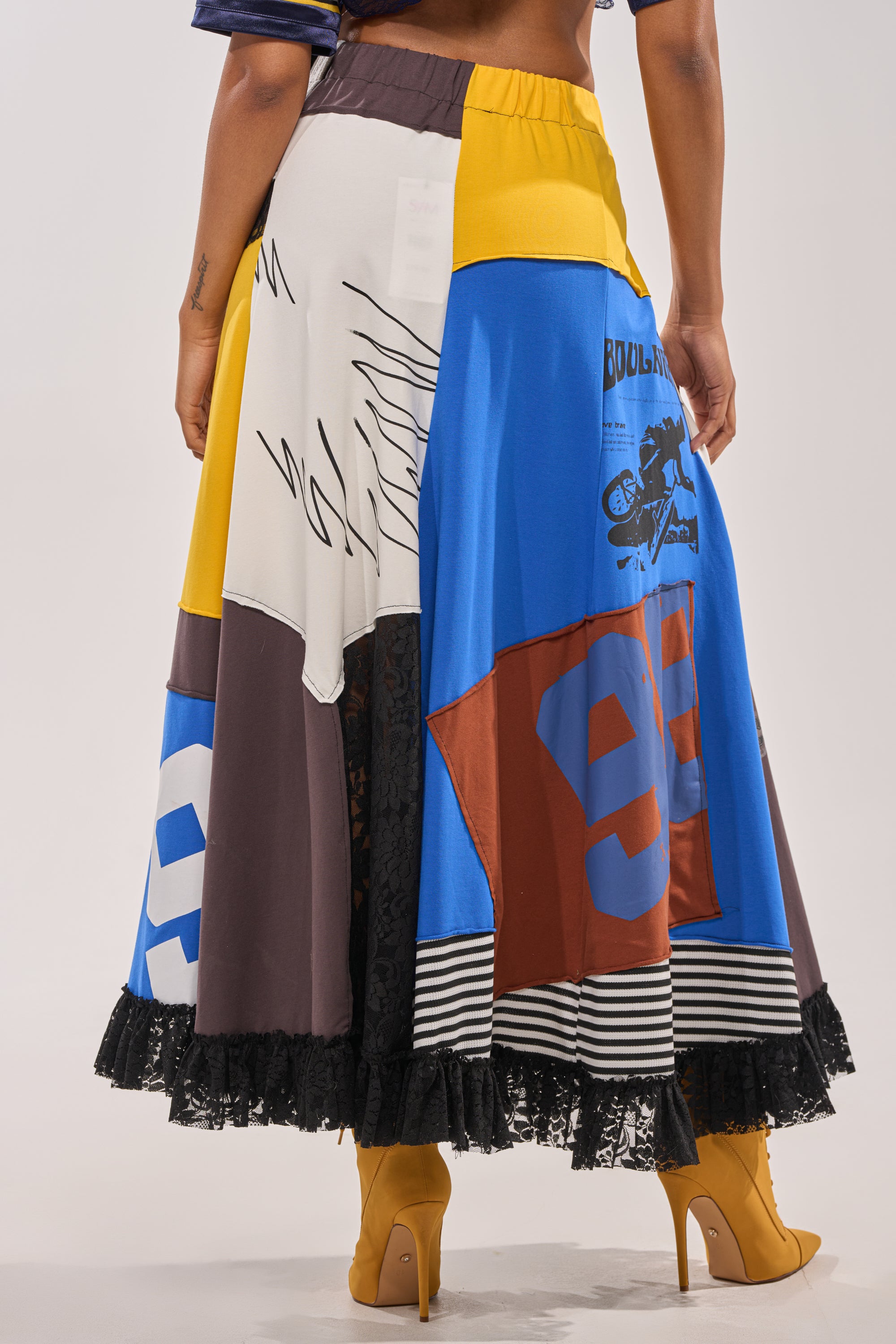 A person photographed from behind wears the CRAZY FOR YOU MAXI SKIRT—a colorful patchwork maxi with graphic prints, yellow accents, and a black lace hem—paired with mustard yellow high-heeled boots. The shot highlights the skirt’s unique details. 5