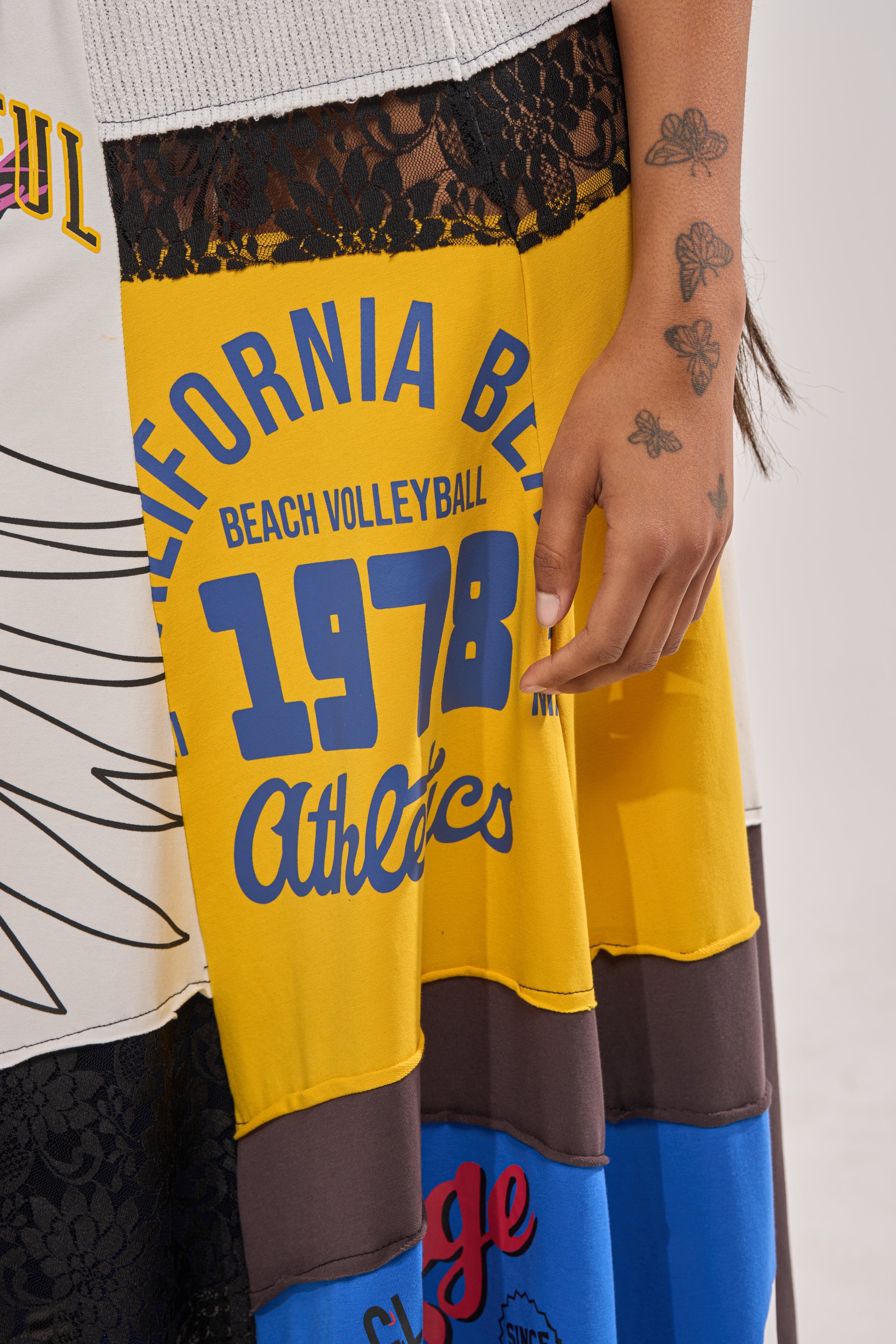 A person with several butterfly tattoos on their hand is wearing the CRAZY FOR YOU MAXI SKIRT, which features black lace, graphic patterns, and the text "California Beach Volleyball 1978 Athletics" in blue and yellow. 7
