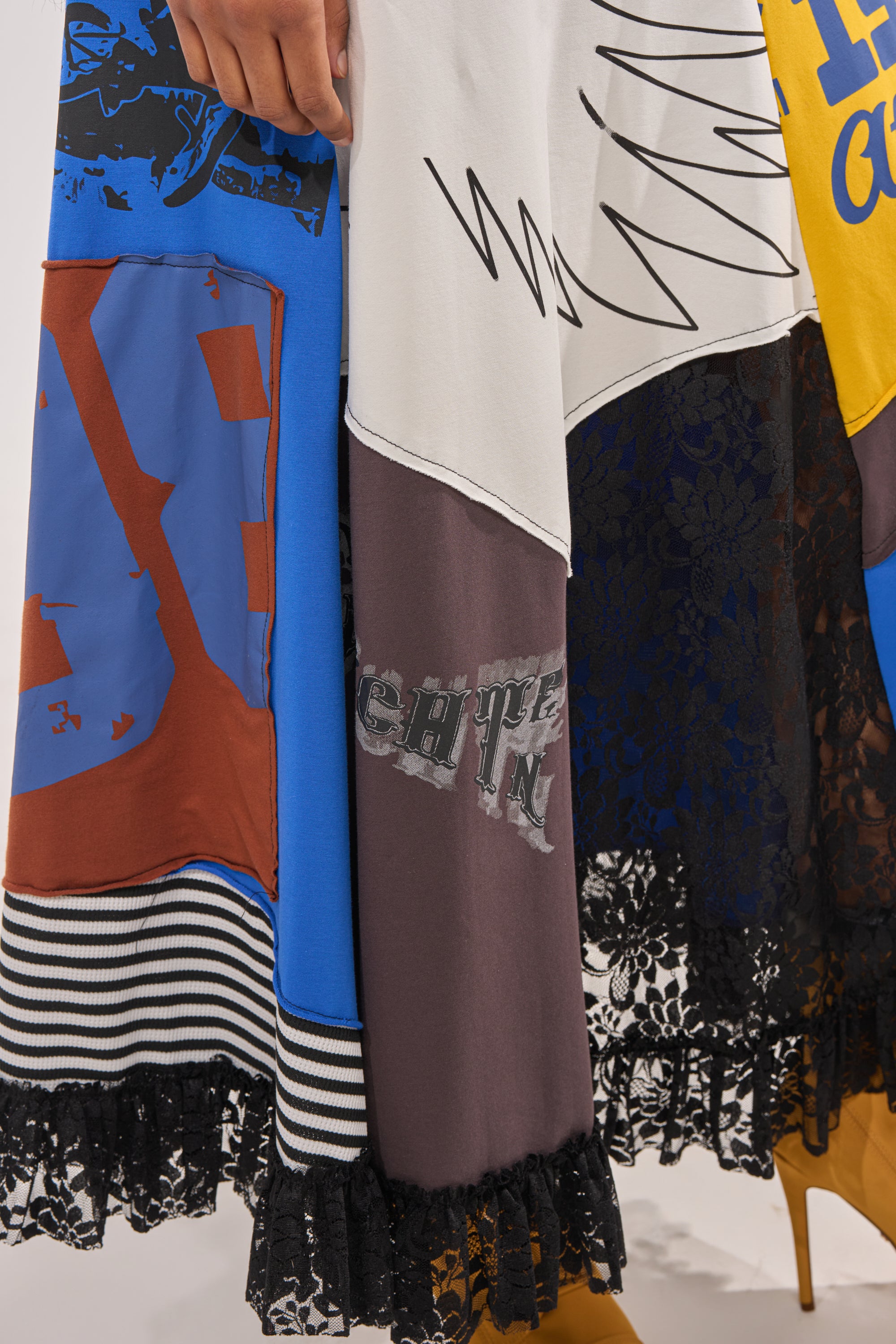 A close-up of the CRAZY FOR YOU MAXI SKIRT shows its vibrant patchwork, stripes, lace trim, and bold graphic patterns. A hand gently holds the skirt as brown boots peek out below. 8