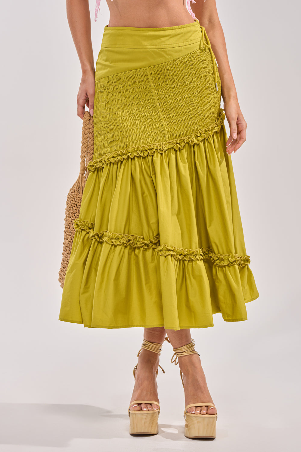 A woman showcases the IT ONLY GETS BETTER RUCHED MAXI SKIRT in vibrant chartreuse, paired with platform sandals and a woven straw bag. The waist-down shot captures effortless resort style. 1