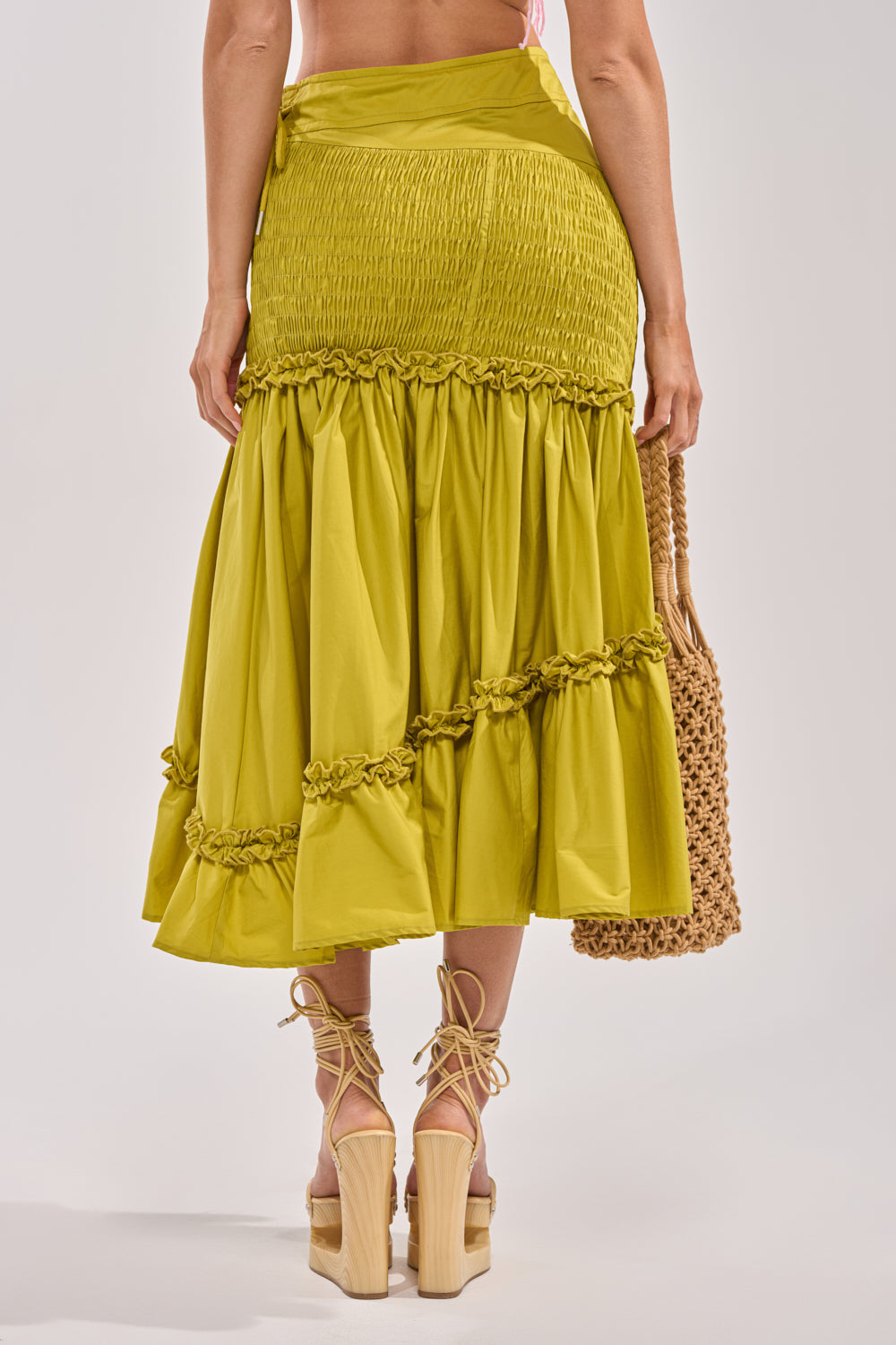 A woman faces away, wearing the IT ONLY GETS BETTER RUCHED MAXI SKIRT in chartreuse green with tiered, gathered details. She holds a woven bag and wears platform sandals with ankle-wrap ties against a plain light background. 3