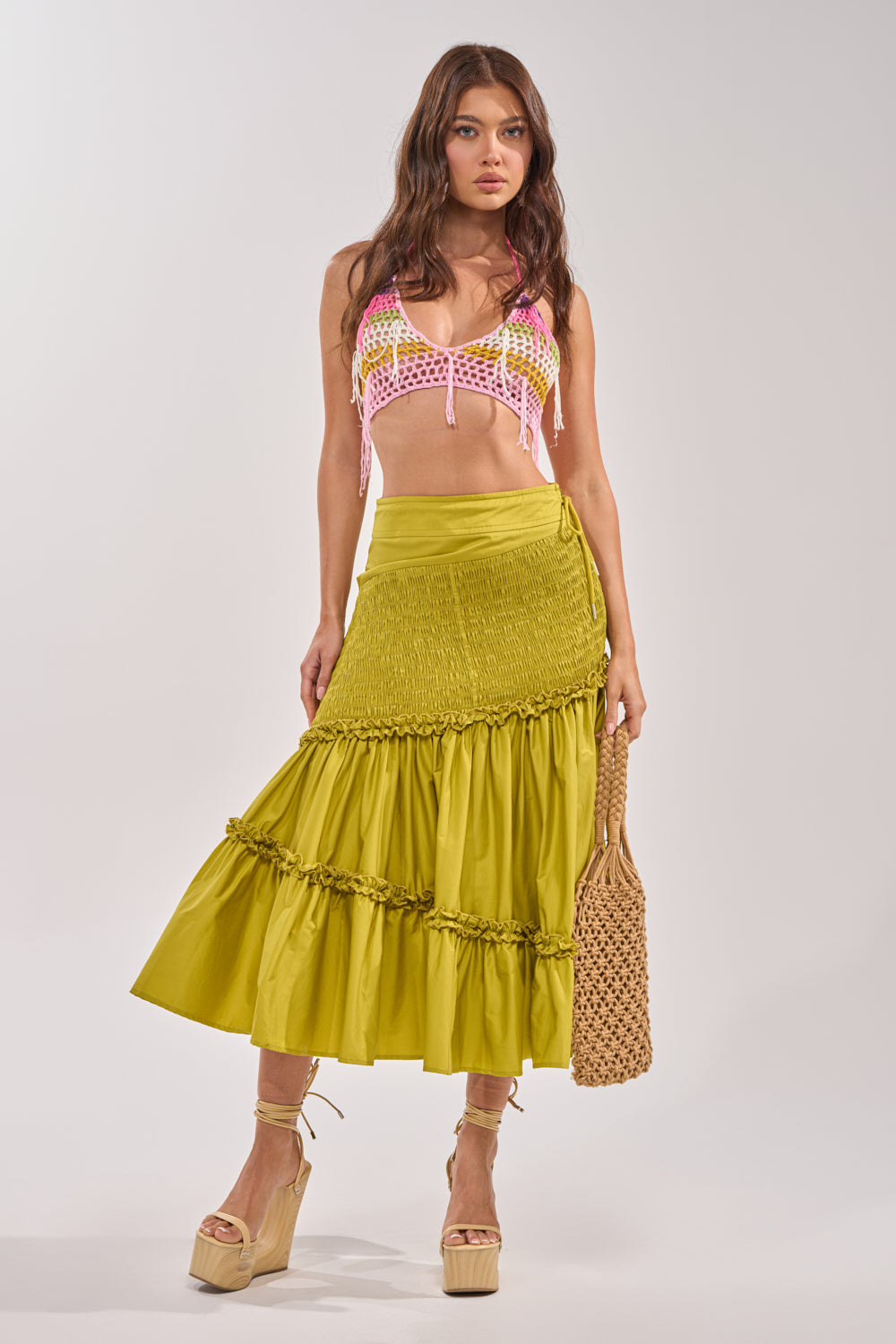 A woman wearing a pink and white crochet crop top, the IT ONLY GETS BETTER RUCHED MAXI SKIRT in chartreuse, tan platform sandals, and holding a woven straw bag stands against a plain white background. 4