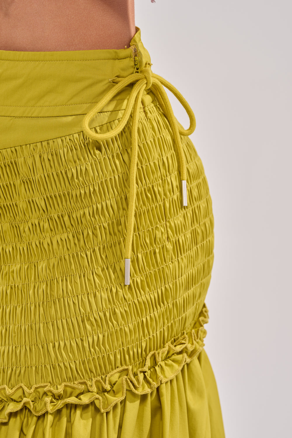 Close-up of the IT ONLY GETS BETTER RUCHED MAXI SKIRT in chartreuse, featuring ruched fabric, elastic textured panels, a drawstring waist with metal tips, and a ruffled hem. Shown against a plain neutral background. 6