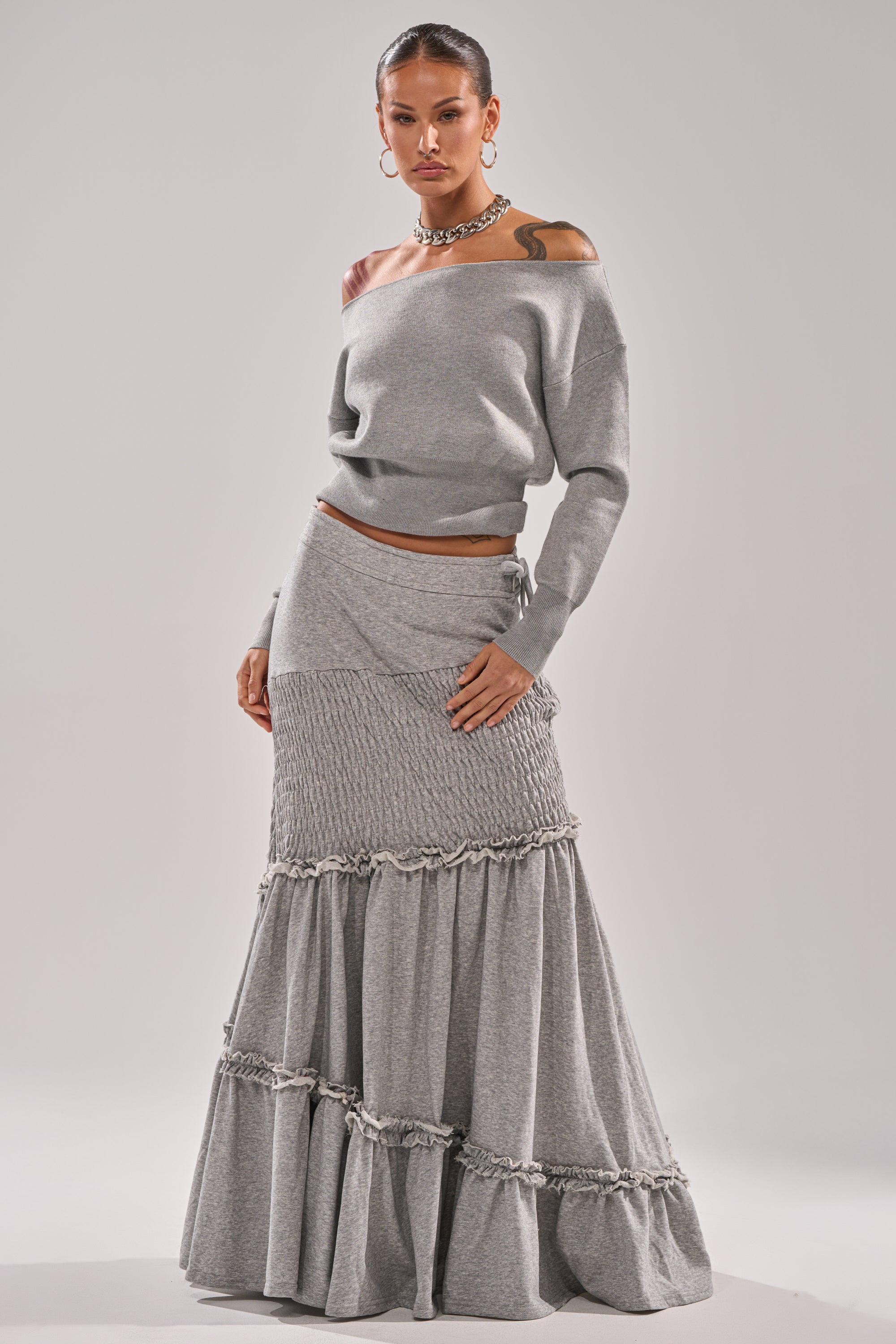 A woman stands confidently in a grey, off-the-shoulder top and the IT ONLY GETS BETTER RUCHED MAXI SKIRT IN HEATHER GREY, styled with hoop earrings and a chunky necklace against a plain background. 0