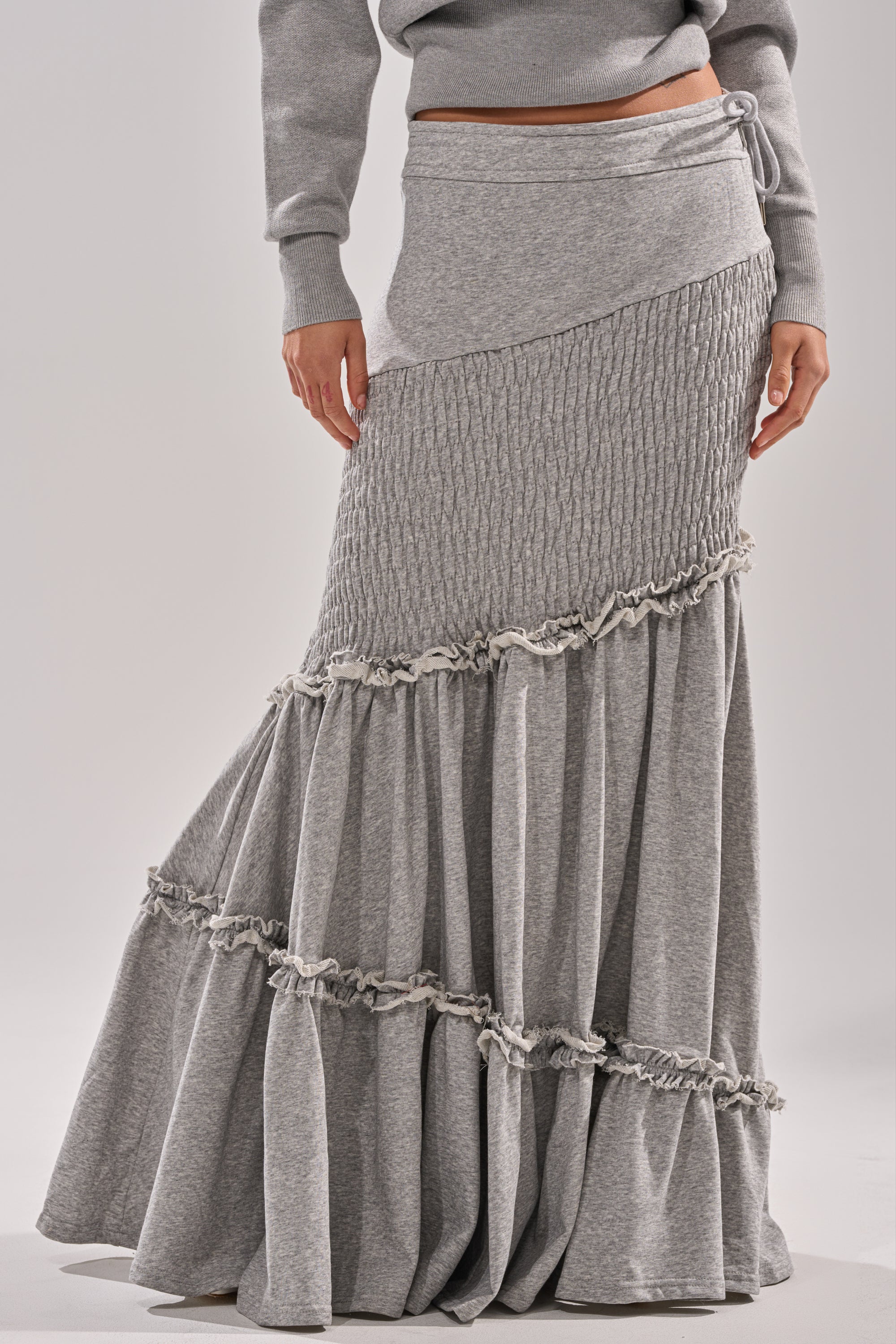 A person models the IT ONLY GETS BETTER RUCHED MAXI SKIRT IN HEATHER GREY—crafted from cotton-blend jersey with smocked panels and ruffle details—styled with a matching grey sweater, set against a plain background. 2