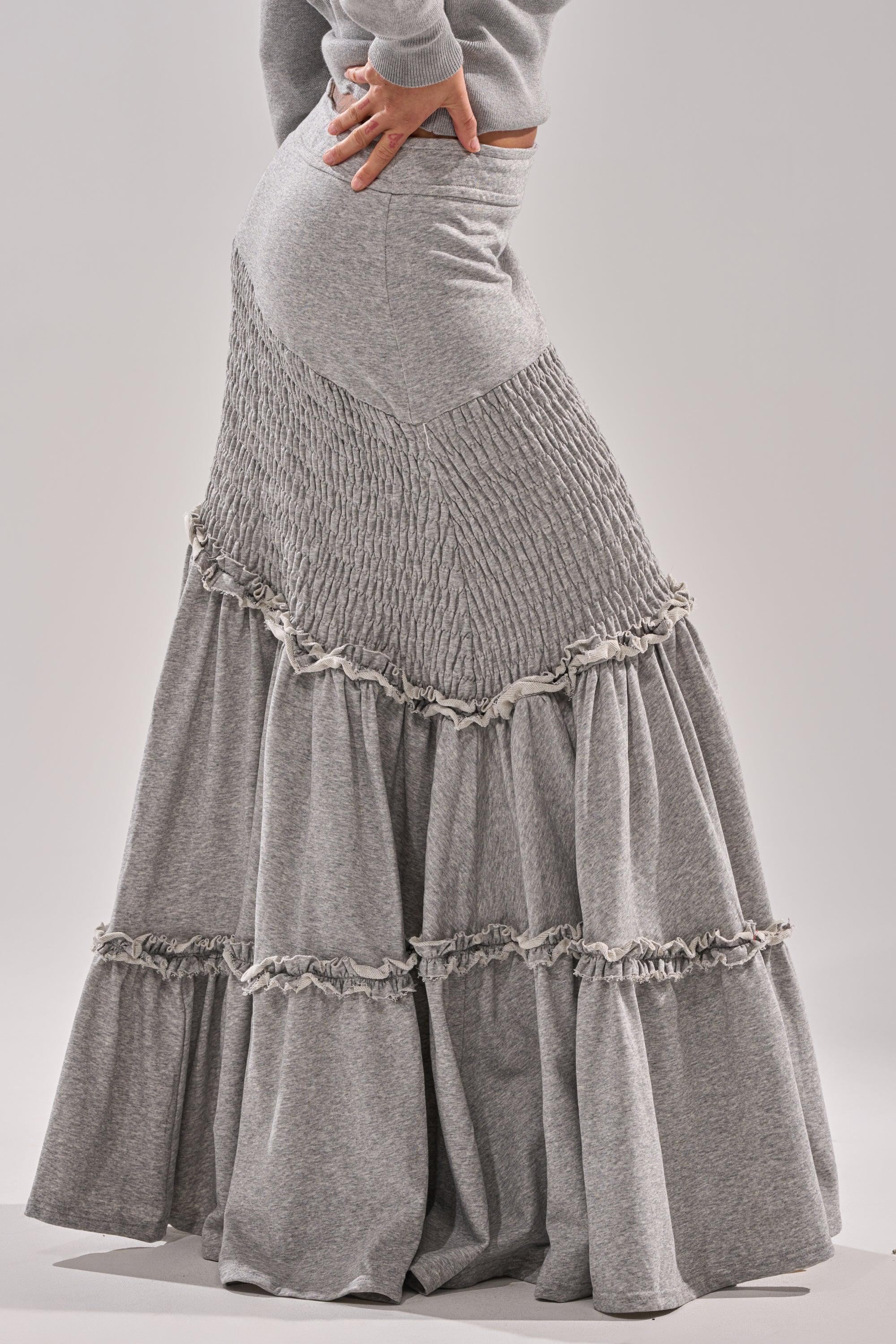 A person poses with one hand on their hip against a plain background, wearing the IT ONLY GETS BETTER RUCHED MAXI SKIRT IN HEATHER GREY, featuring gathered and ruffled tiered details. 3