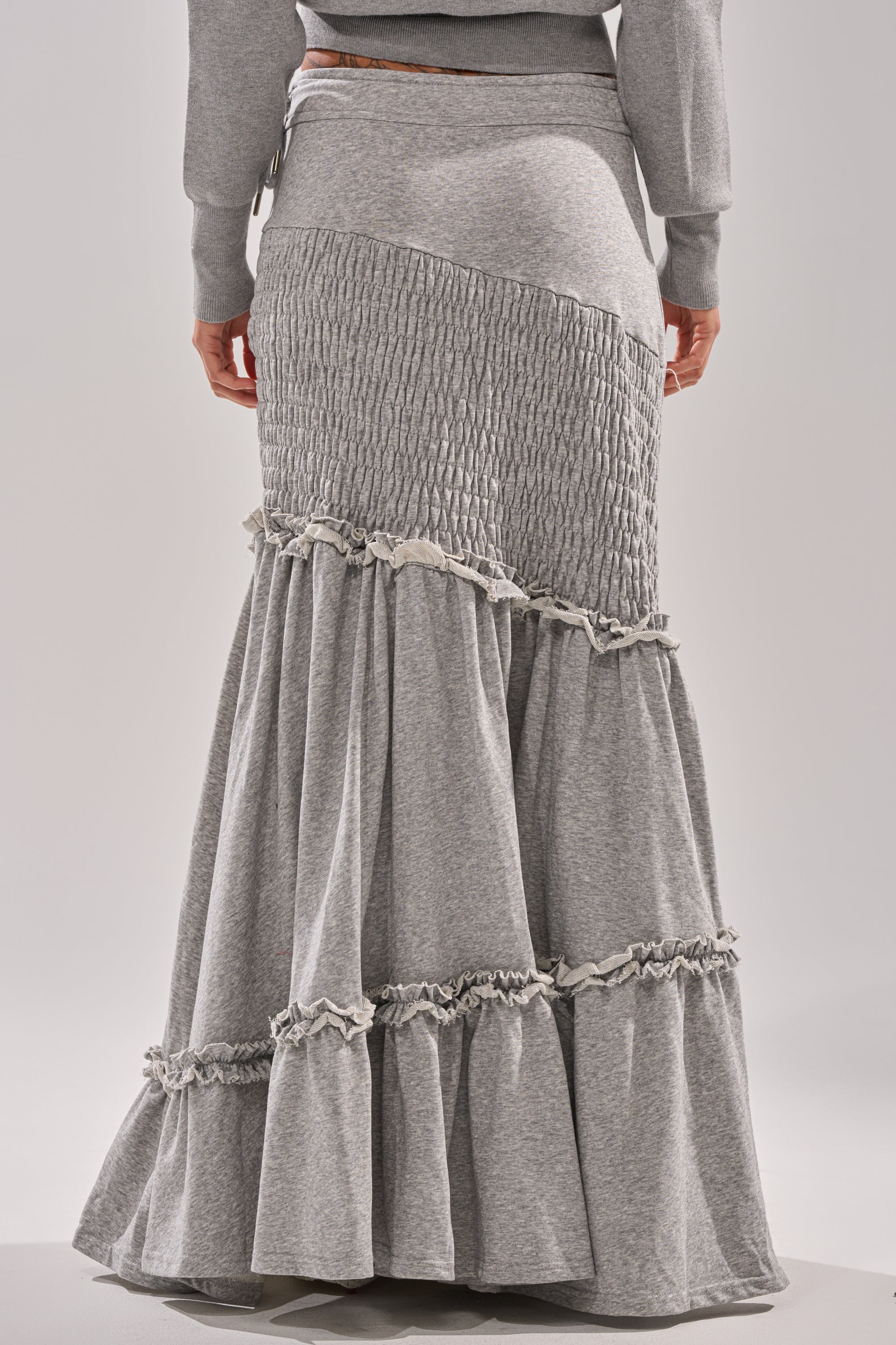 A person wears the IT ONLY GETS BETTER RUCHED MAXI SKIRT IN HEATHER GREY, featuring textured, ruched panels and ruffled seams for a layered look. The back view image shows the skirt from the lower torso to the feet. 4