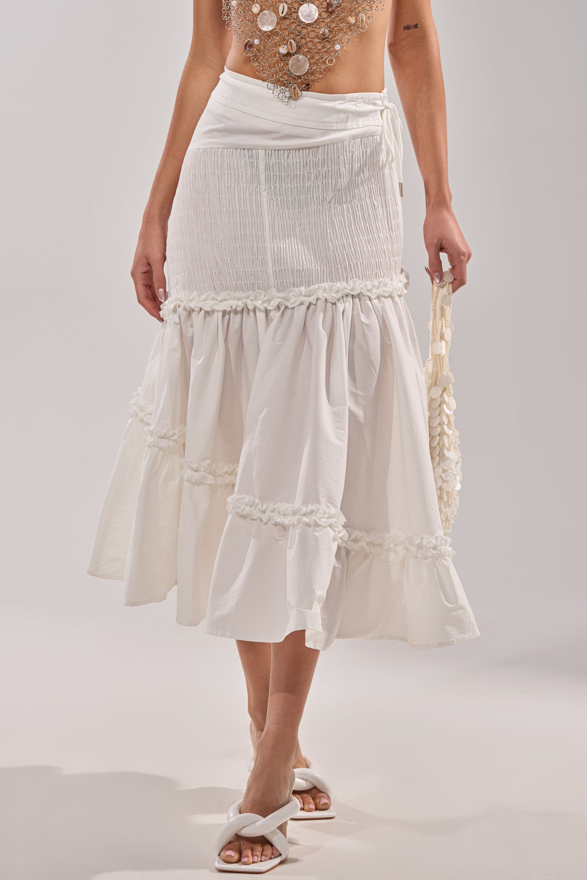 A woman styles THE BEST RUCHED SKIRT—a white, tiered midi with ruffle trim and a smocked high-rise waist—with white sandals, a woven bag, and a shell-detailed top peeking into view.