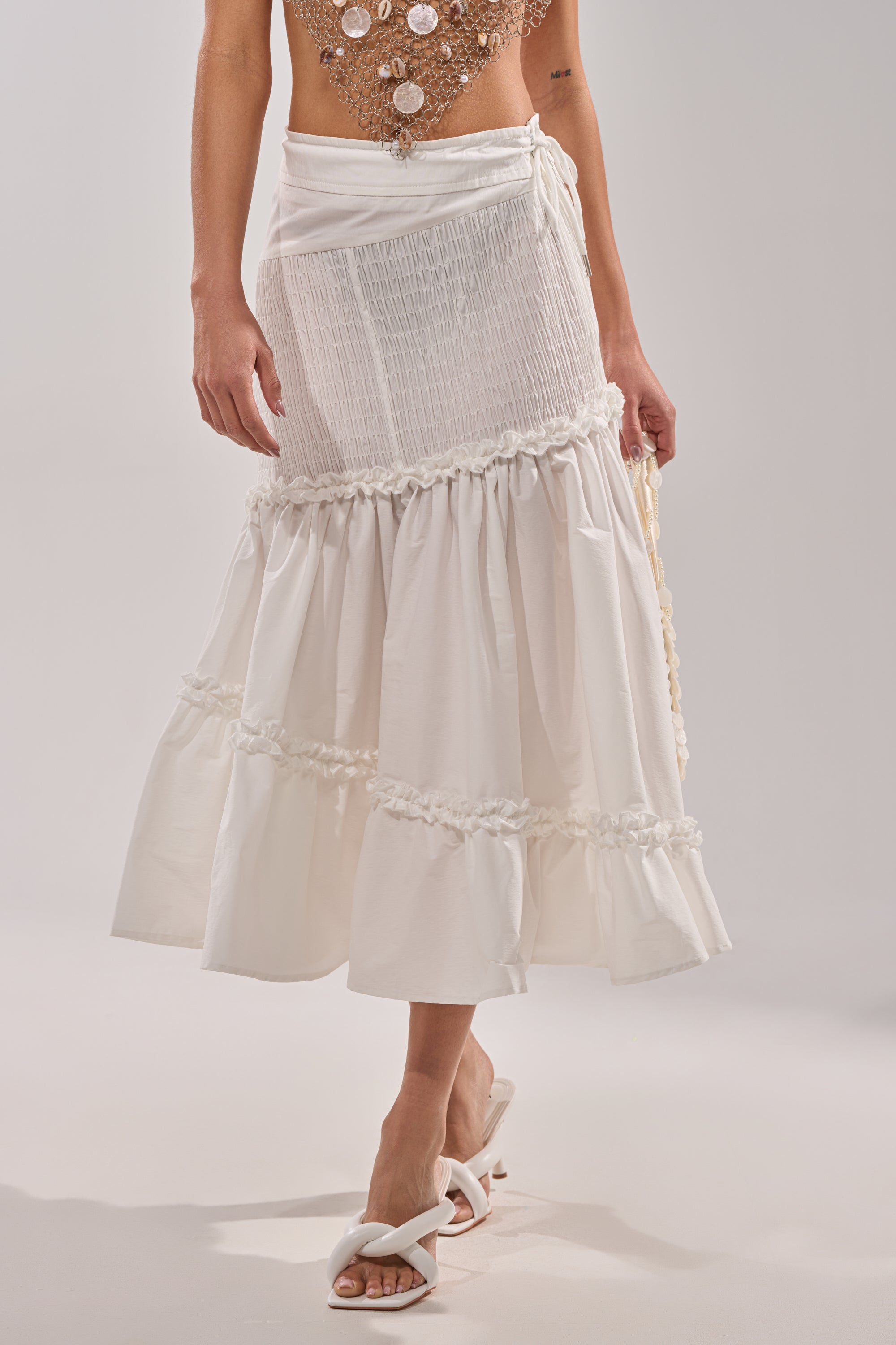 Someone is wearing THE BEST RUCHED SKIRT, a white, ankle-length ruffle trim skirt with gathered details, styled with white heeled sandals. Only the lower body in this tiered midi skirt appears against a plain light background.
