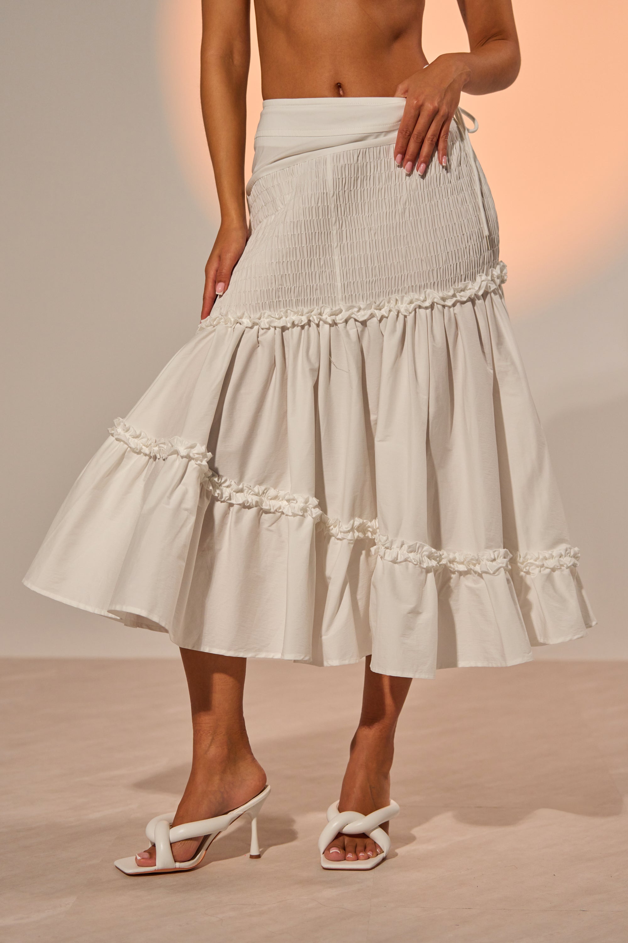 A woman models THE BEST RUCHED SKIRT, a white high-waisted, tiered midi with ruffle trim and gathered details, paired with white heeled sandals. Only her midsection and legs are shown against a neutral background.