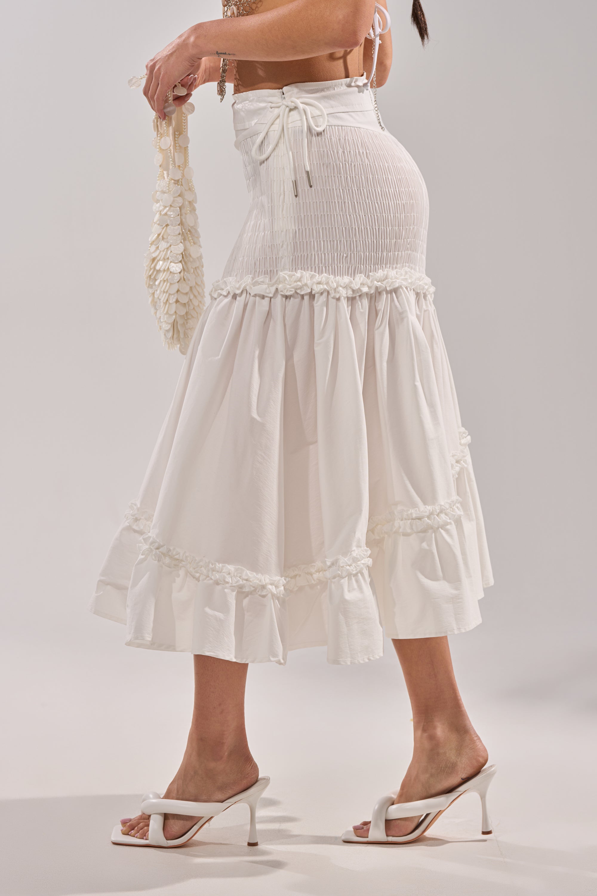 A woman wears THE BEST RUCHED SKIRT with white heeled sandals and holds a cream textured handbag, standing against a plain light background.