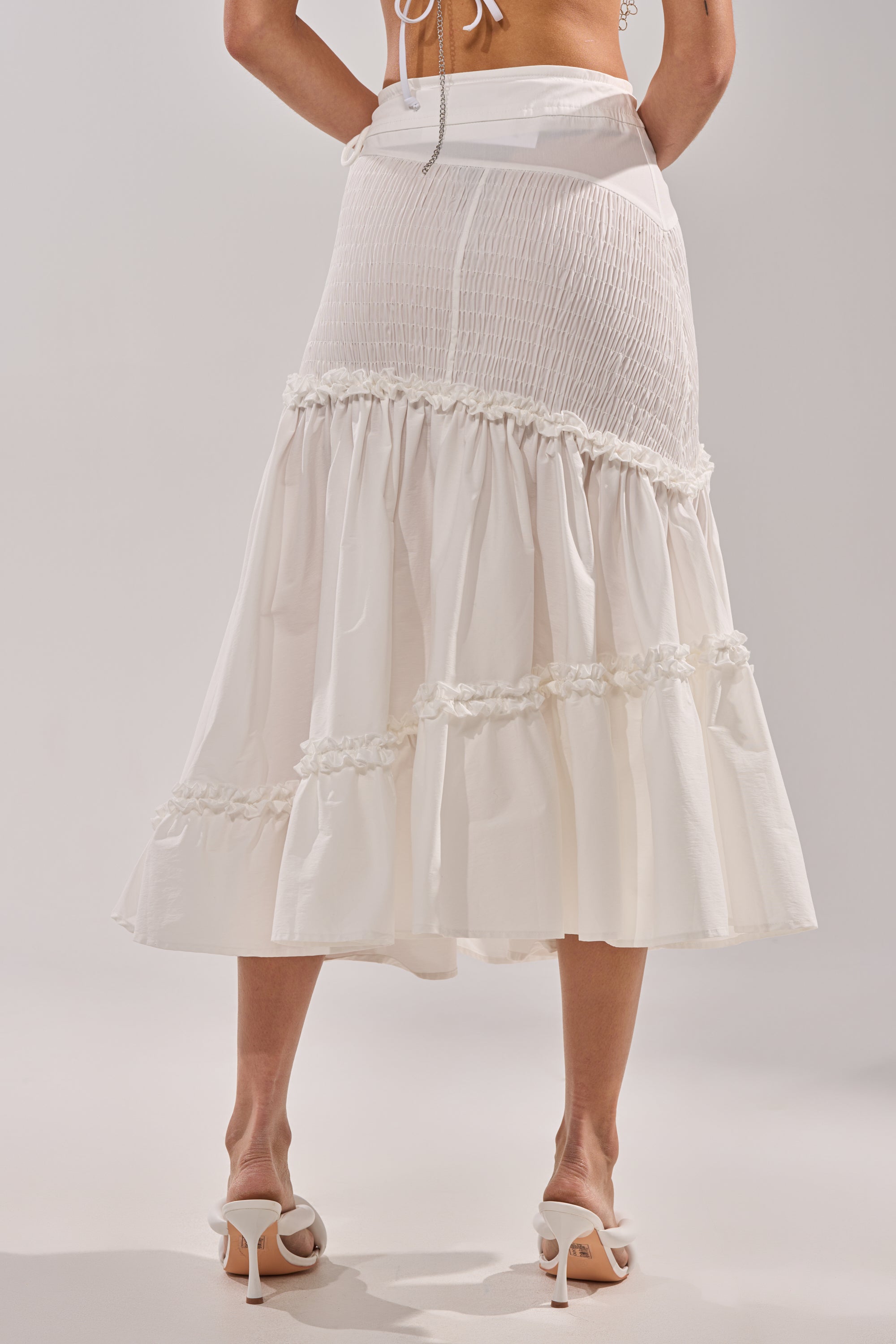 A woman is photographed from the back against a plain light background, wearing THE BEST RUCHED SKIRT in white with tiered ruffle trim and white heeled sandals.