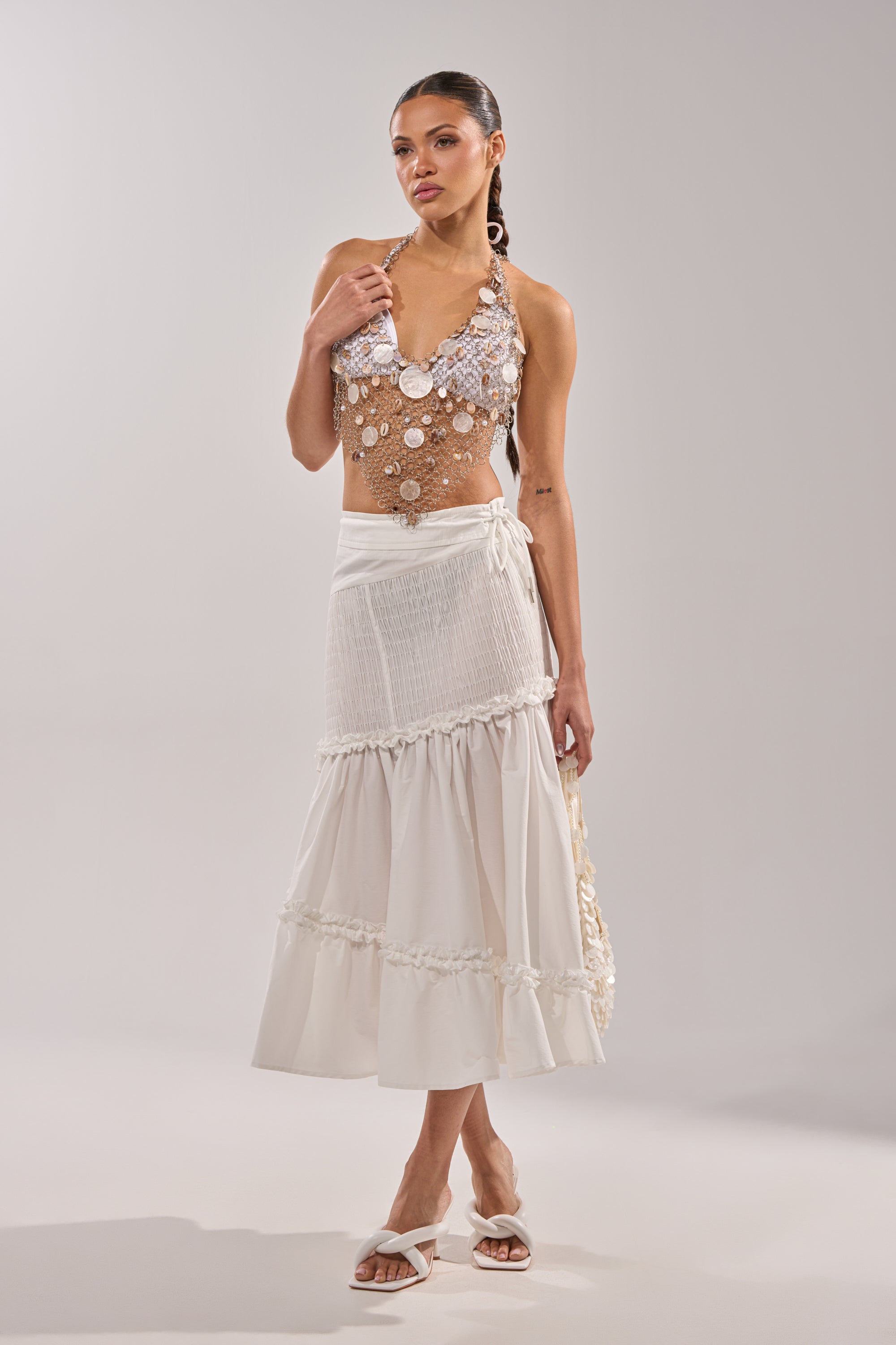 A woman poses in THE BEST RUCHED SKIRT—a white, tiered midi skirt with ruffle trim—paired with a sequined halter top and white heeled sandals against a light background.