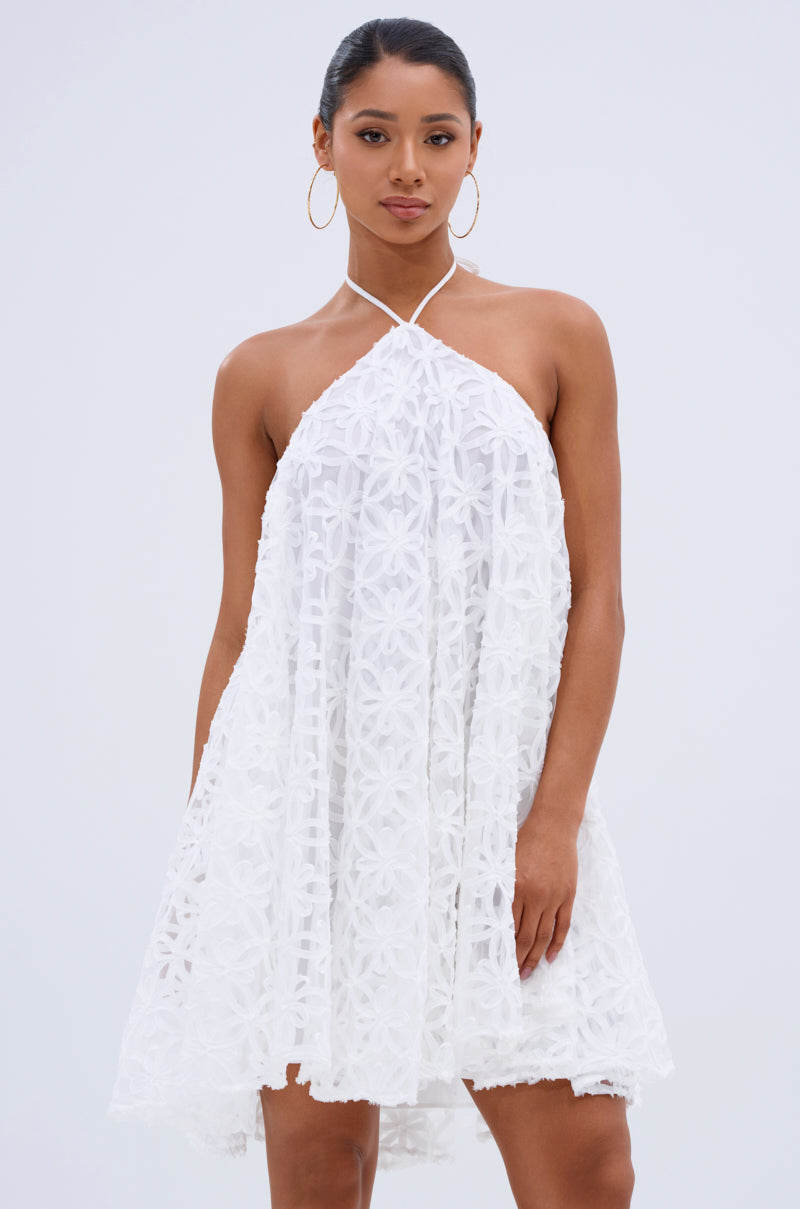 A woman wears the LIZA HALTER MINI DRESS, featuring delicate floral detailing, and poses confidently against a plain light background. 0