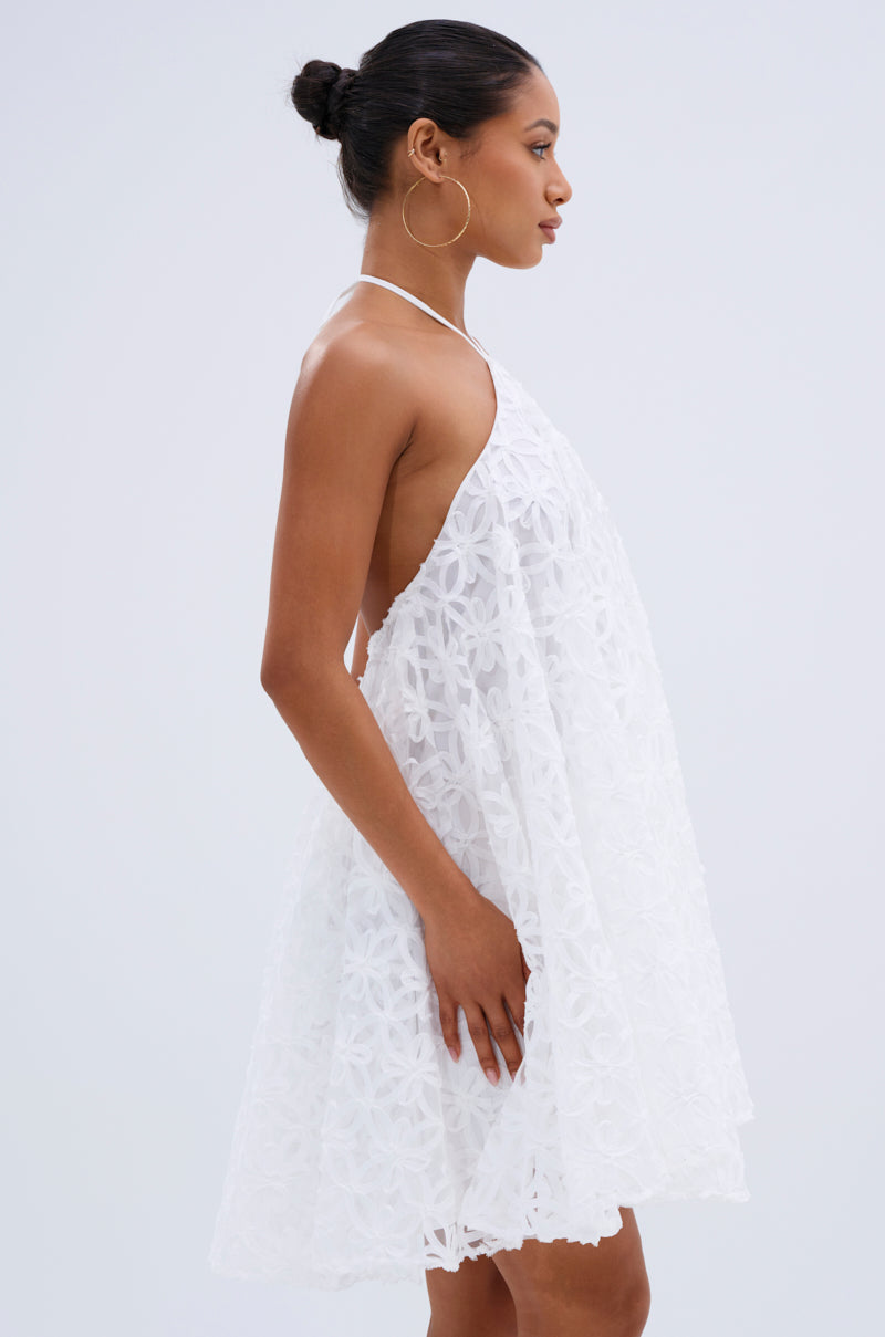 A woman with her hair in a bun, wearing large hoop earrings and the LIZA HALTER MINI DRESS with elegant floral lace detailing, stands in profile against a plain light background. 1