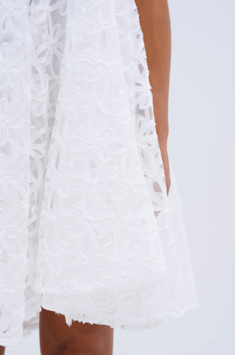 Close-up of a person wearing the LIZA HALTER MINI DRESS, a white mini dress with textured 3D floral details and a flared skirt. Part of their brown arm and leg is visible against a light background. 4
