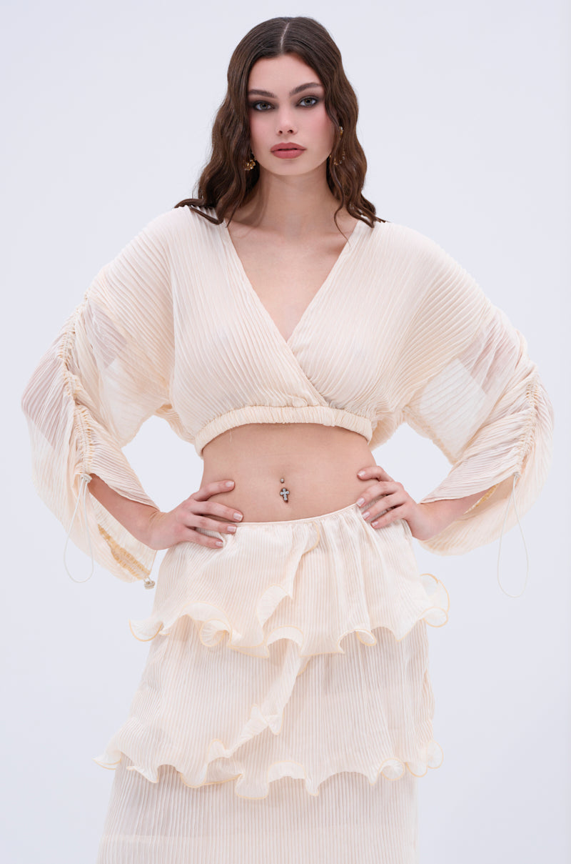 A woman with wavy brown hair stands hands on hips against a plain background, wearing the sheer, cream-colored RUFFLE HEAVEN BLOUSE featuring a cropped silhouette, wide sleeves, and a matching tiered skirt.