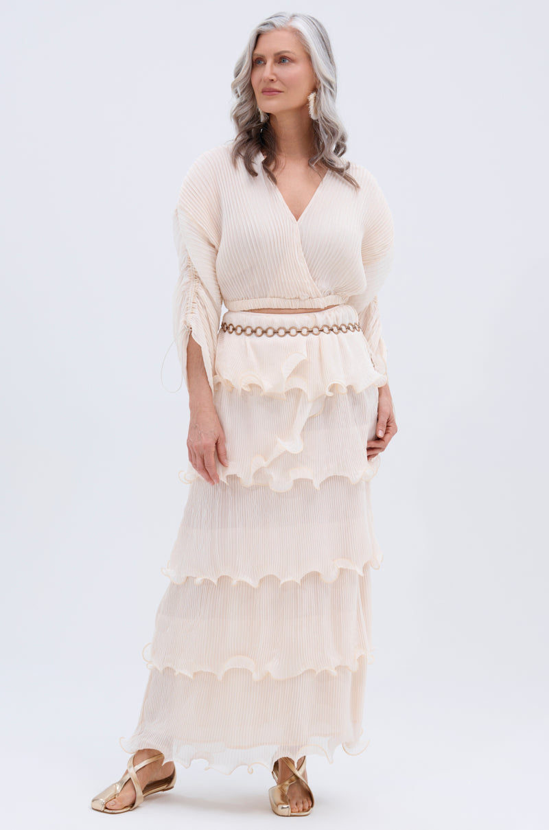A woman with long gray hair wears the RUFFLE HEAVEN BLOUSE, a cream-colored top with ruffled sheer layers and a decorative belt, paired with gold sandals, standing against a plain white background.