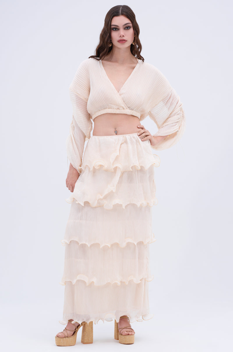 A woman stands against a white background in a cream, long-sleeved crop top and the RUFFLE HEAVEN MAXI SKIRT with tiered ruffles, paired with platform sandals—embodying chic style in every detail.