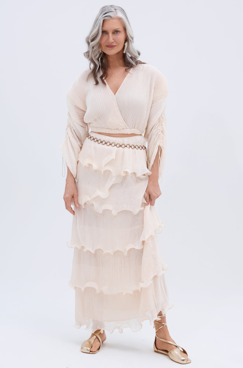 A woman with long, wavy gray hair stands against a white background, wearing a flowing cream RUFFLE HEAVEN MAXI SKIRT with tiered ruffles and a belt with circular links, paired with gold flat sandals.