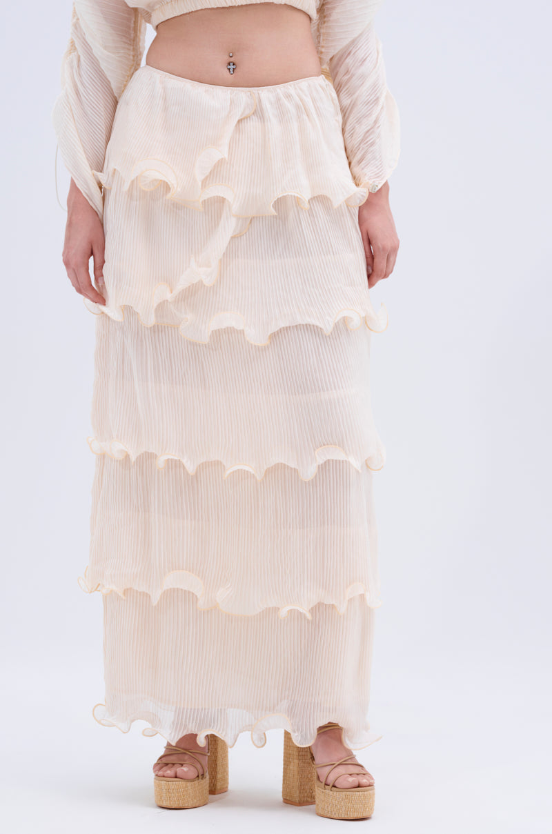 A person models the lower half of the RUFFLE HEAVEN MAXI SKIRT, a cream-colored, sheer maxi with tiered ruffles, paired with a matching crop top and tan platform sandals, shown against a plain light background.