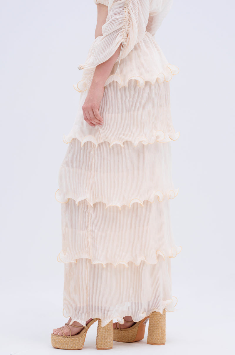 A person wears the RUFFLE HEAVEN MAXI SKIRT, a sheer, pleated, cream-colored skirt with tiered ruffles and platform sandals. The image is cropped at the shoulders, highlighting the skirt's texture against a plain background.