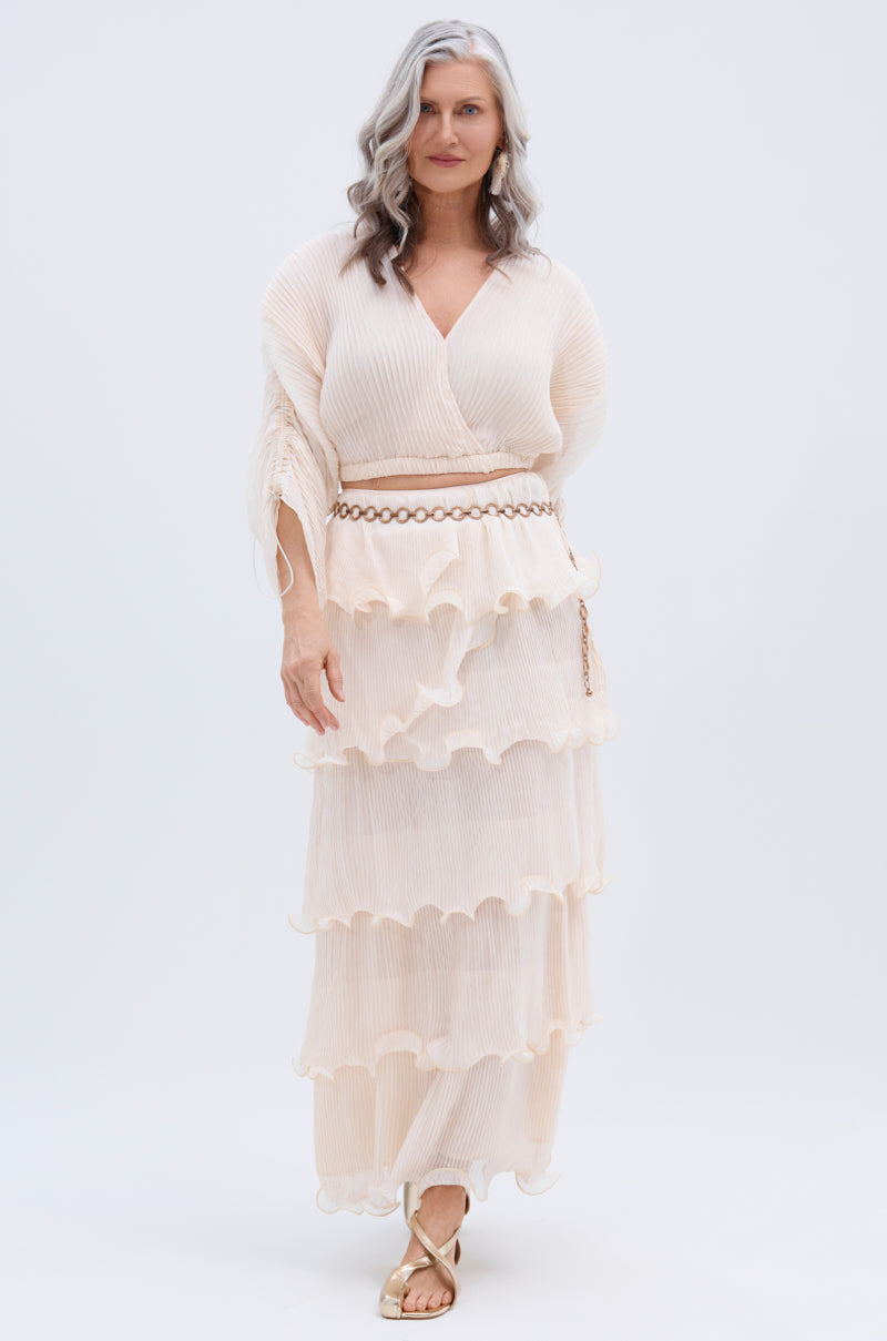 A woman with long, wavy gray hair wears the RUFFLE HEAVEN MAXI SKIRT—a cream-colored, tiered ruffle skirt—styled with sheer sleeves, a chain belt, gold sandals, and set against a plain white background.