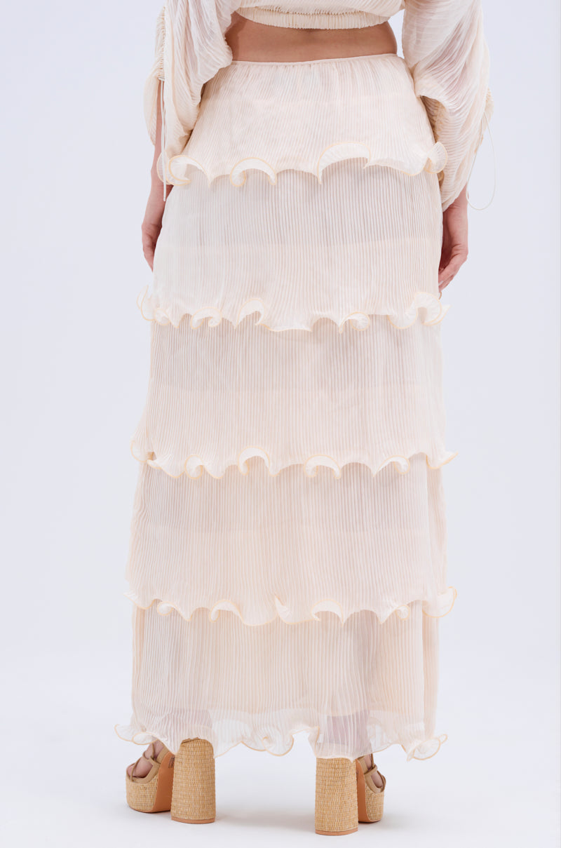 Someone is shown from the back in the RUFFLE HEAVEN MAXI SKIRT—a sheer, light beige maxi with tiered ruffles and vertical pleats—paired with chunky tan platform sandals against a plain light background.