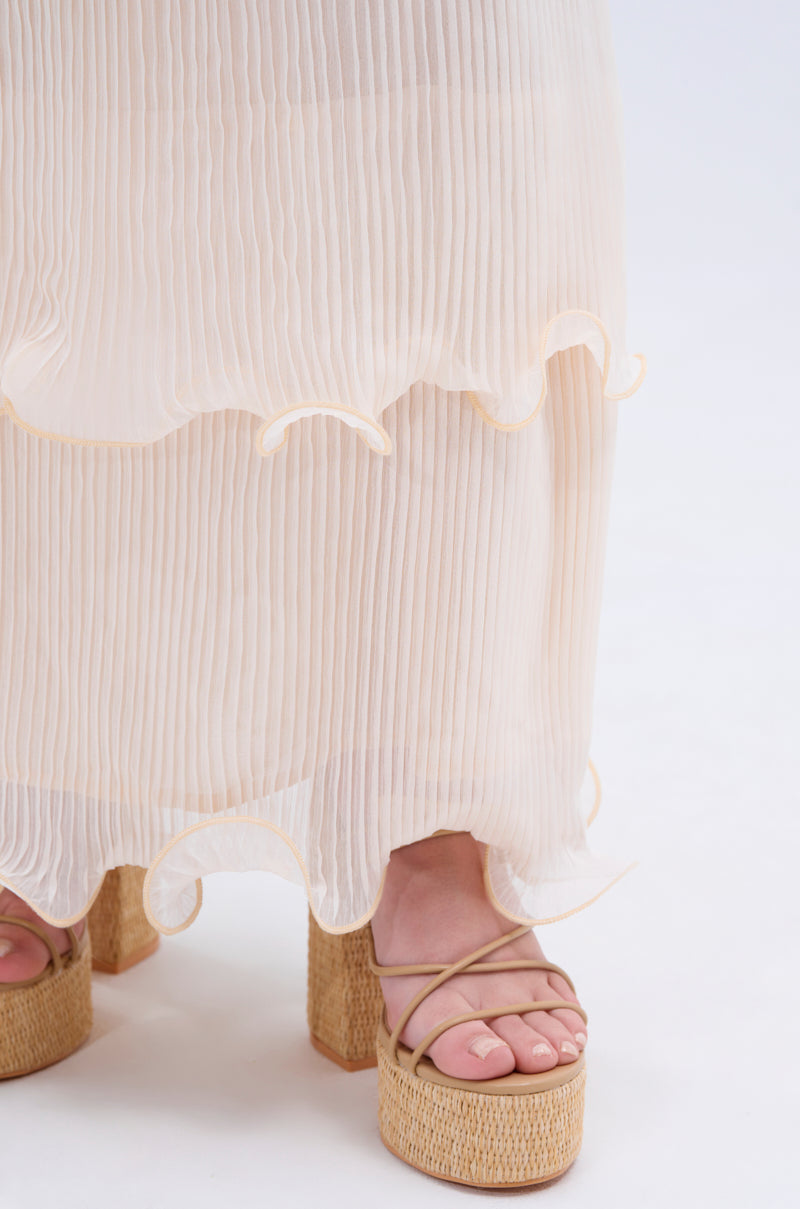 A close-up shows feet in light tan platform sandals with thin straps, partly covered by the RUFFLE HEAVEN MAXI SKIRT—a sheer, pleated maxi skirt with cream ruffled tiers—against a plain, light background for a soft effect.
