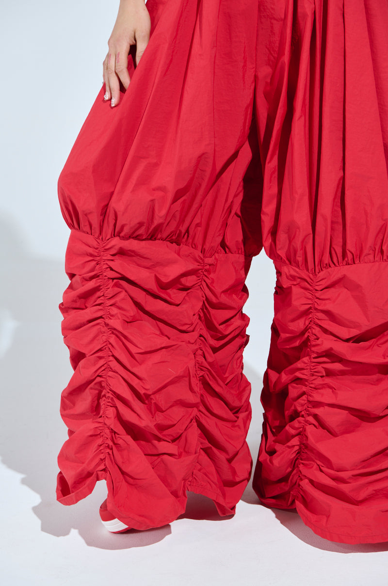 A person wears WIDE LEG SCRUNCHED HEM PANTS in bold red, featuring a high-rise fit and dramatic scrunched hem from the knees down, with their hand resting on one leg against a plain white background.