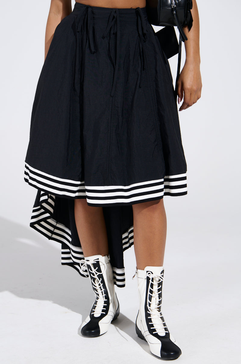 Dressed in the TIME IS UP MIDI SKIRT featuring a unique high-low silhouette and black design with white hem stripes, a person completes the outfit with chic black and white lace-up boots, creating a standout look against the plain white background.