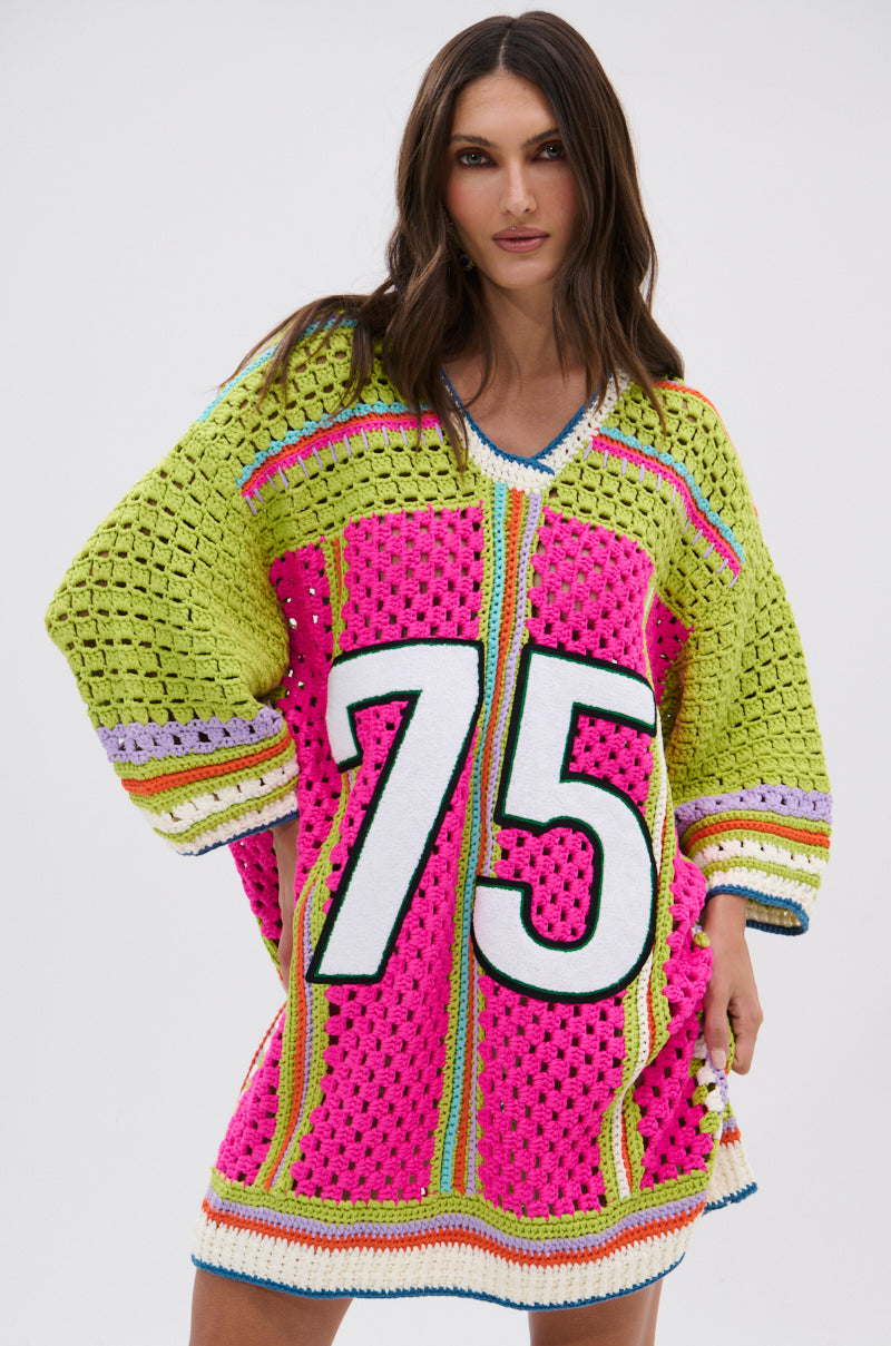 A woman models the COURTSIDE OR SEASIDE CROCHET JERSEY DRESS—a loose, colorful piece with yellow, green, pink, and blue panels and a bold white "75" on the front—radiating sporty charm against a plain background.