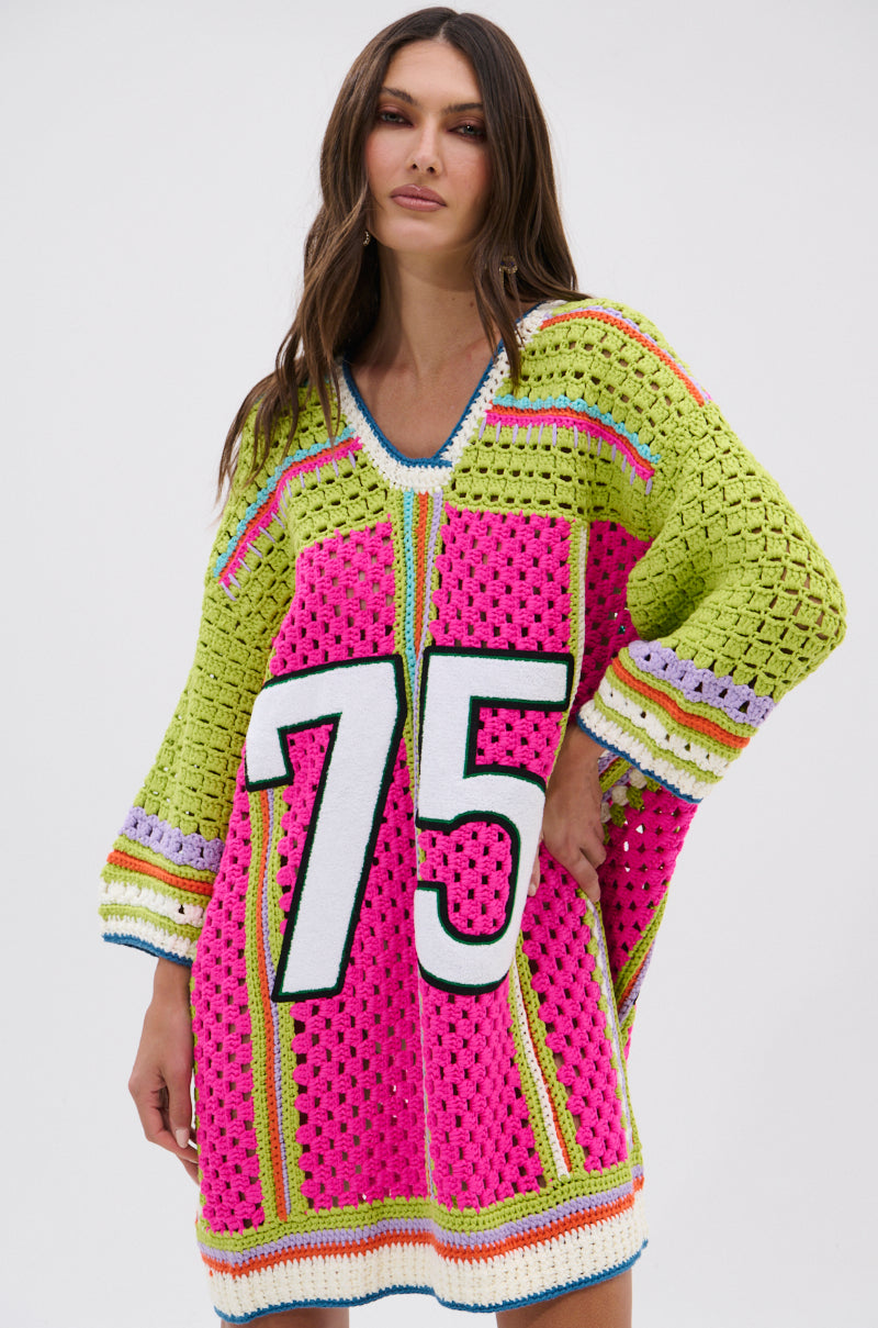 A woman poses against a plain white background wearing the COURTSIDE OR SEASIDE CROCHET JERSEY DRESS, an oversized crochet piece with wide sleeves, a bold white number 75 on the front, and bright color block patterns in pink, green, yellow, and blue.
